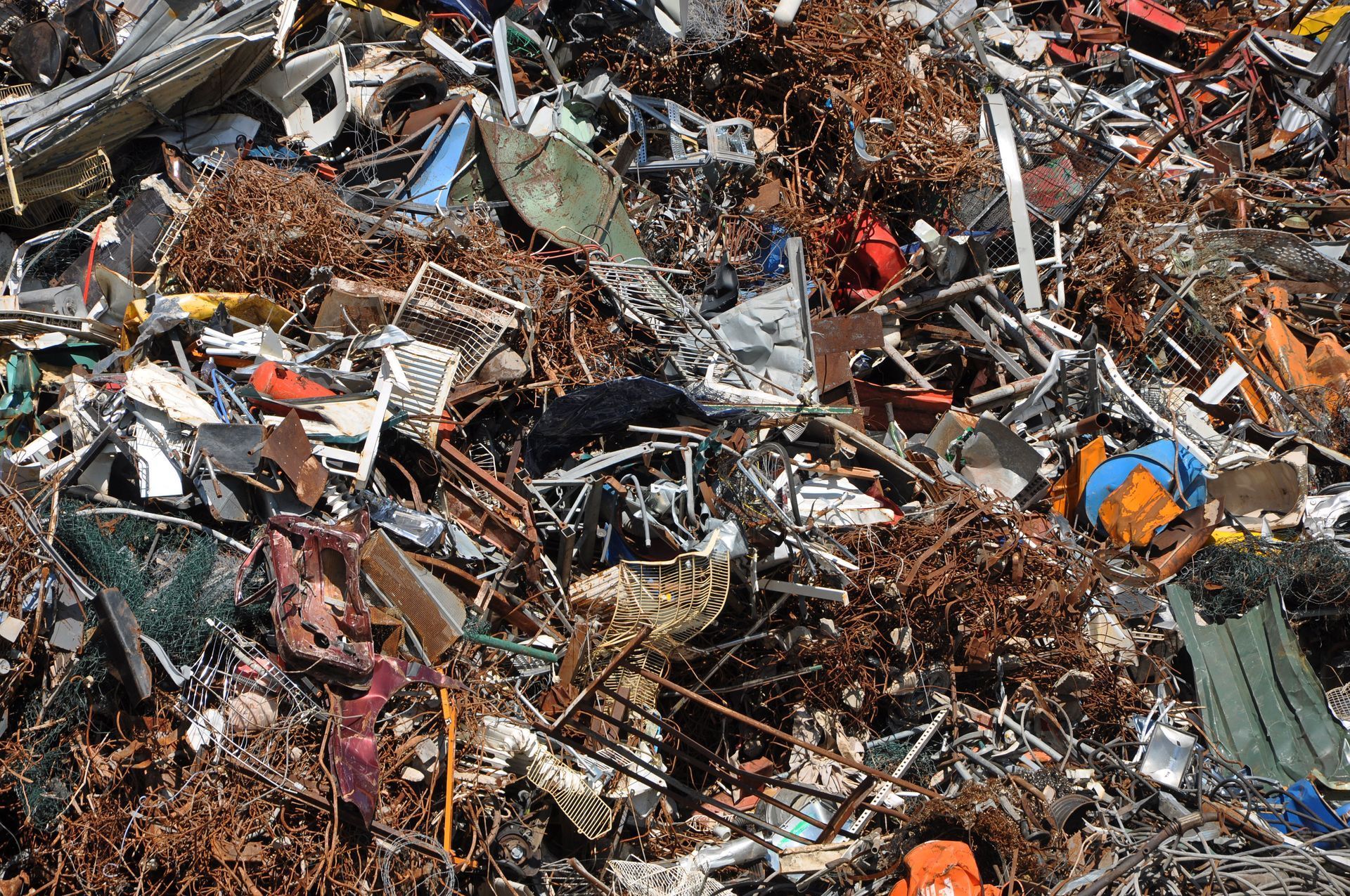Pile of mixed scrap metal and debris in a junkyard, with rusted parts and scattered orange and blue pieces.