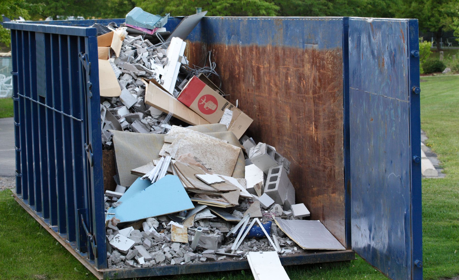 Overflowing blue dumpster filled with mixed construction debris on grass outdoors