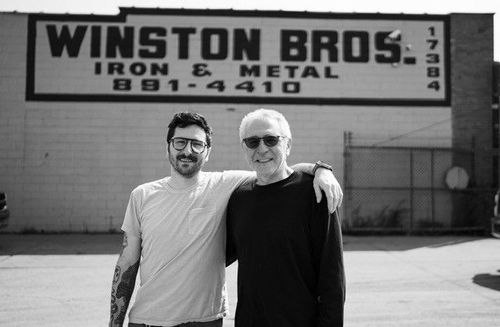 Two people standing arm-in-arm in front of a WINSTON BROS. Iron & Metal sign.