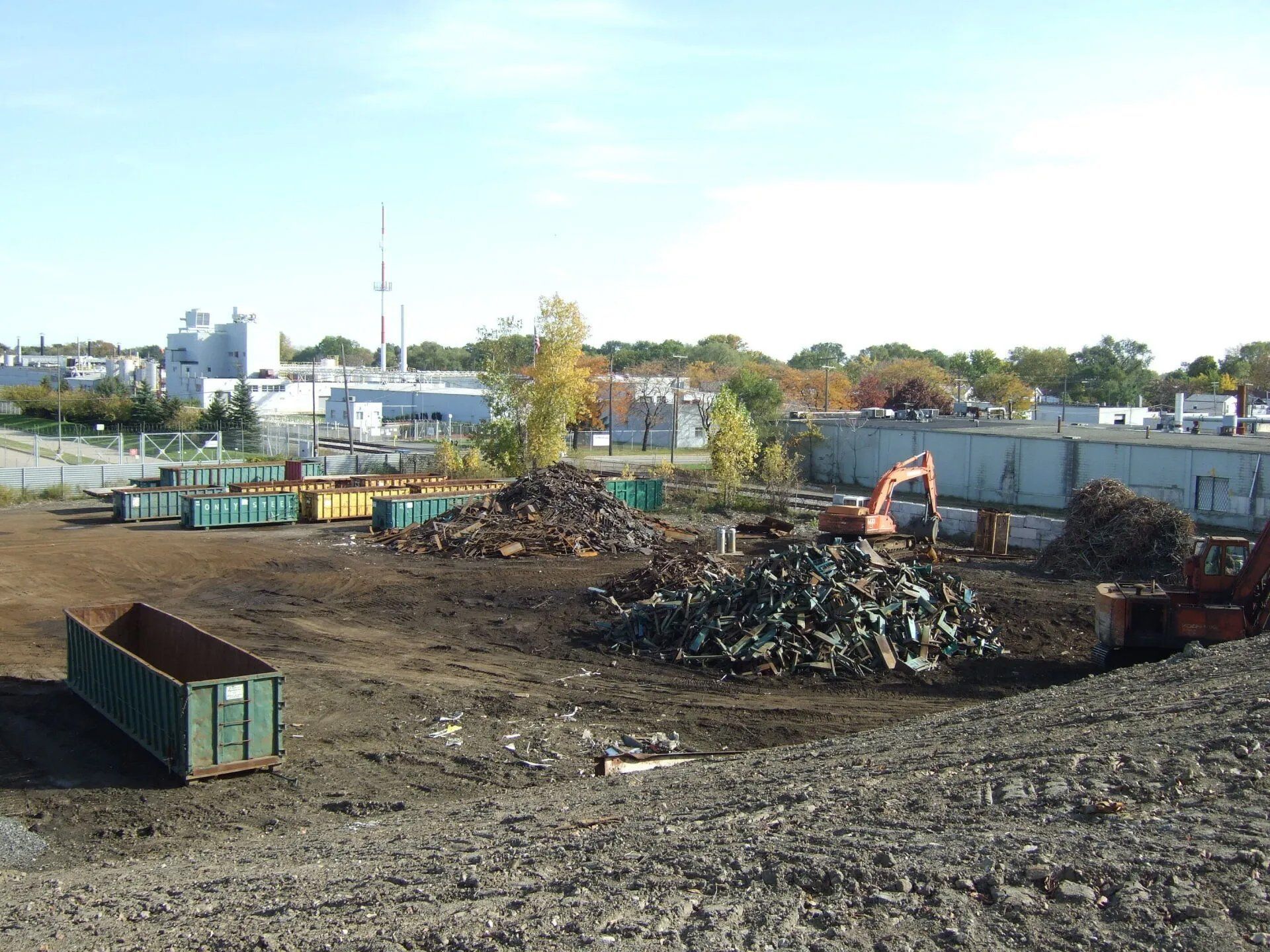 Large dirt lot with scrap piles, dumpsters, and industrial buildings under a clear sky