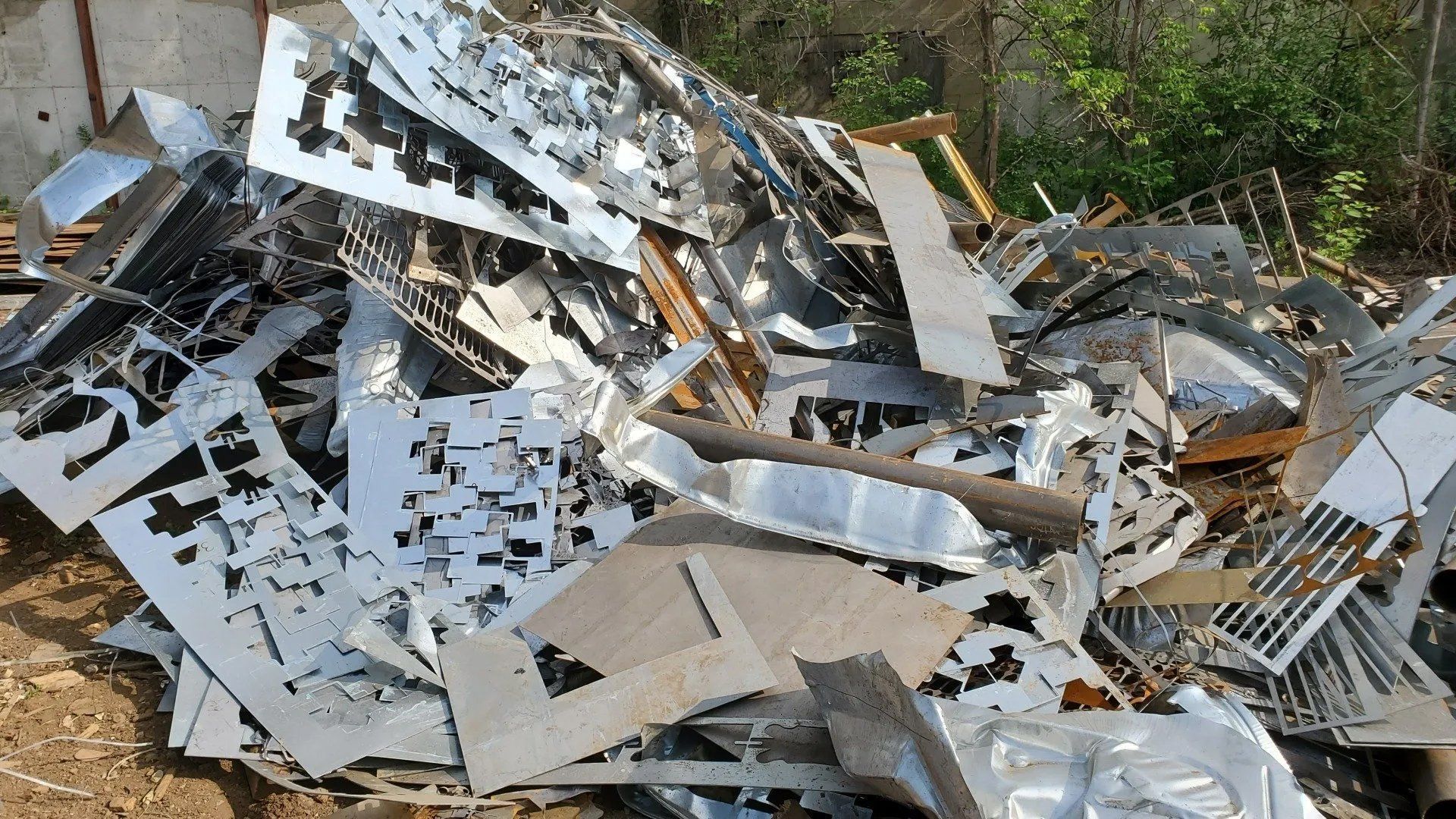 Pile of metal scrap and broken panels scattered outdoors near trees and a fence.