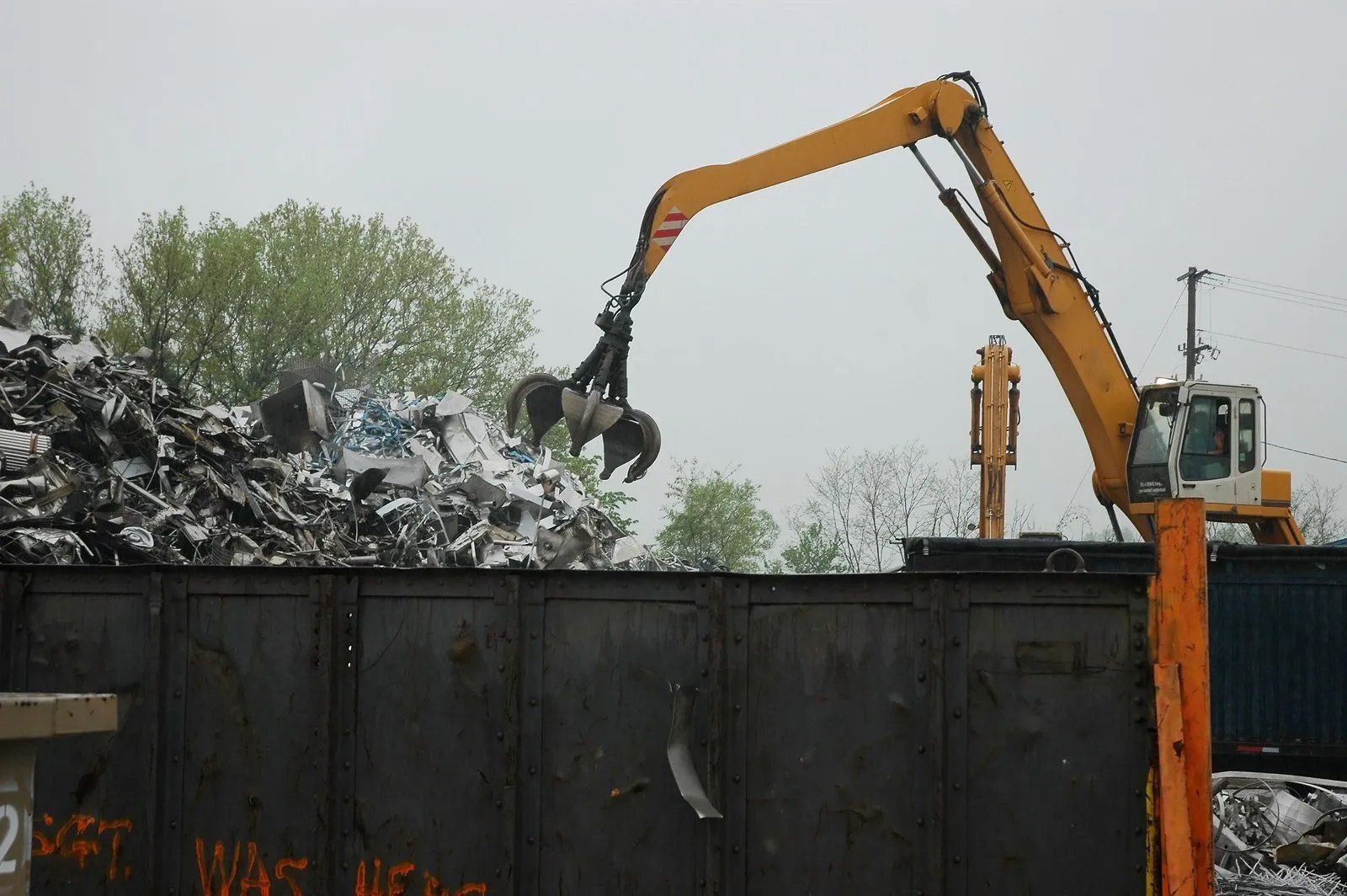 Yellow excavator loading scrap metal into a large container at a recycling yard.