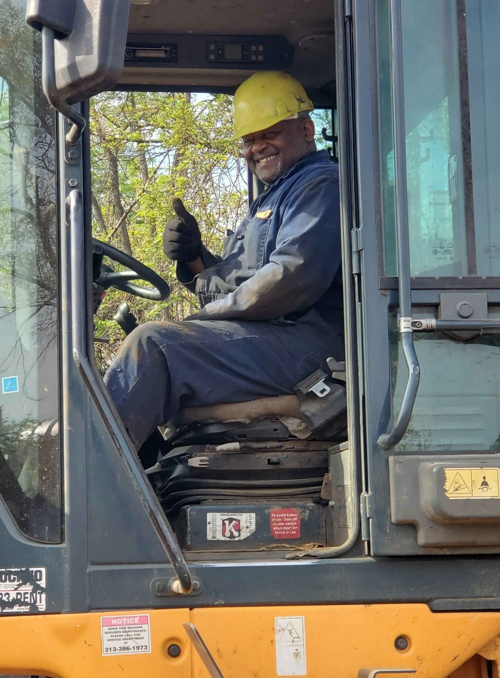 Construction worker in yellow hard hat sitting in an excavator cab, giving a thumbs-up.