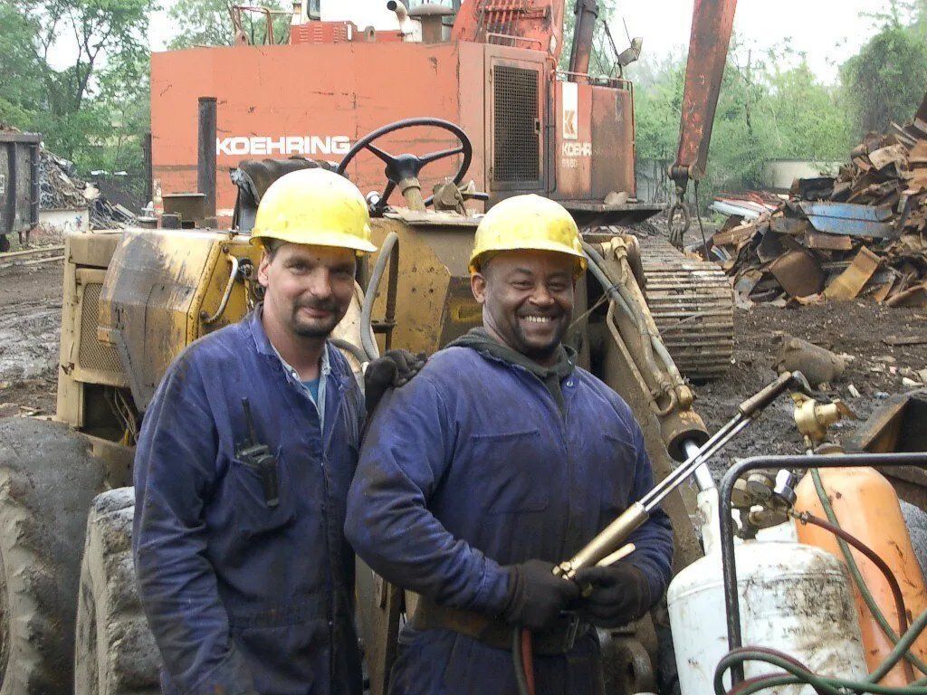 Two workers in hard hats stand by heavy machinery at a construction site, smiling.