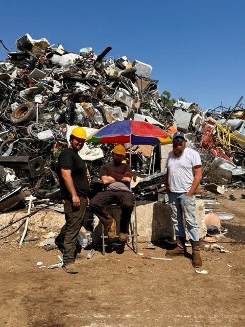 Three people stand beside a cart under a red-blue umbrella in a scrapyard of piled metal.