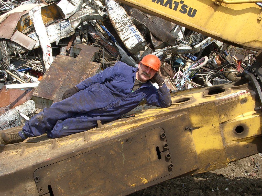 Worker in orange hard hat lying on a yellow excavator amid a scrap metal pile.