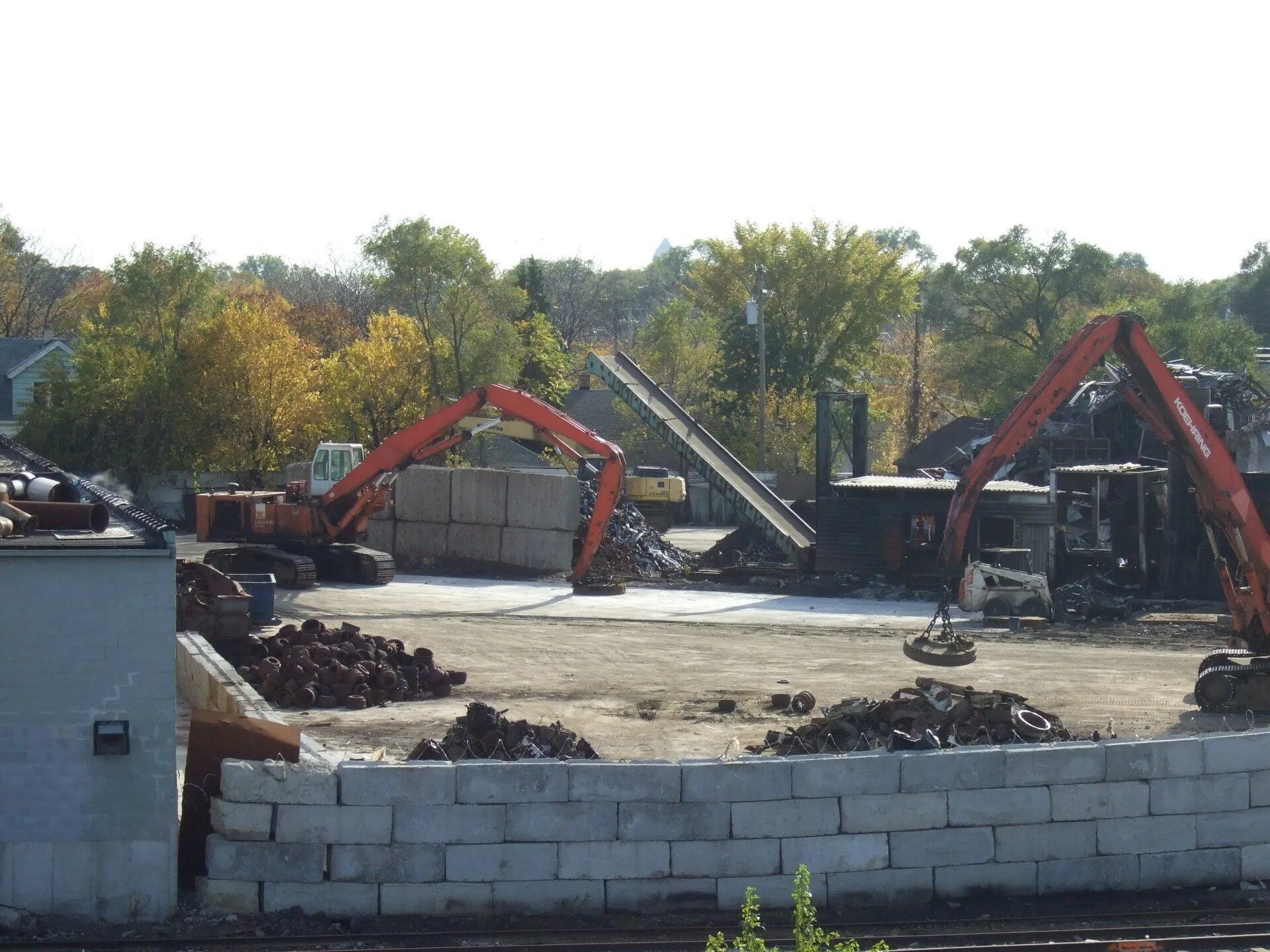 Construction site with red excavators, debris piles, and a concrete block wall under clear sky