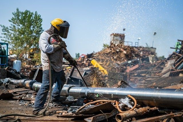 Worker in yellow hard hat uses a shovel at a scrapyard with piles of metal debris under a clear sky