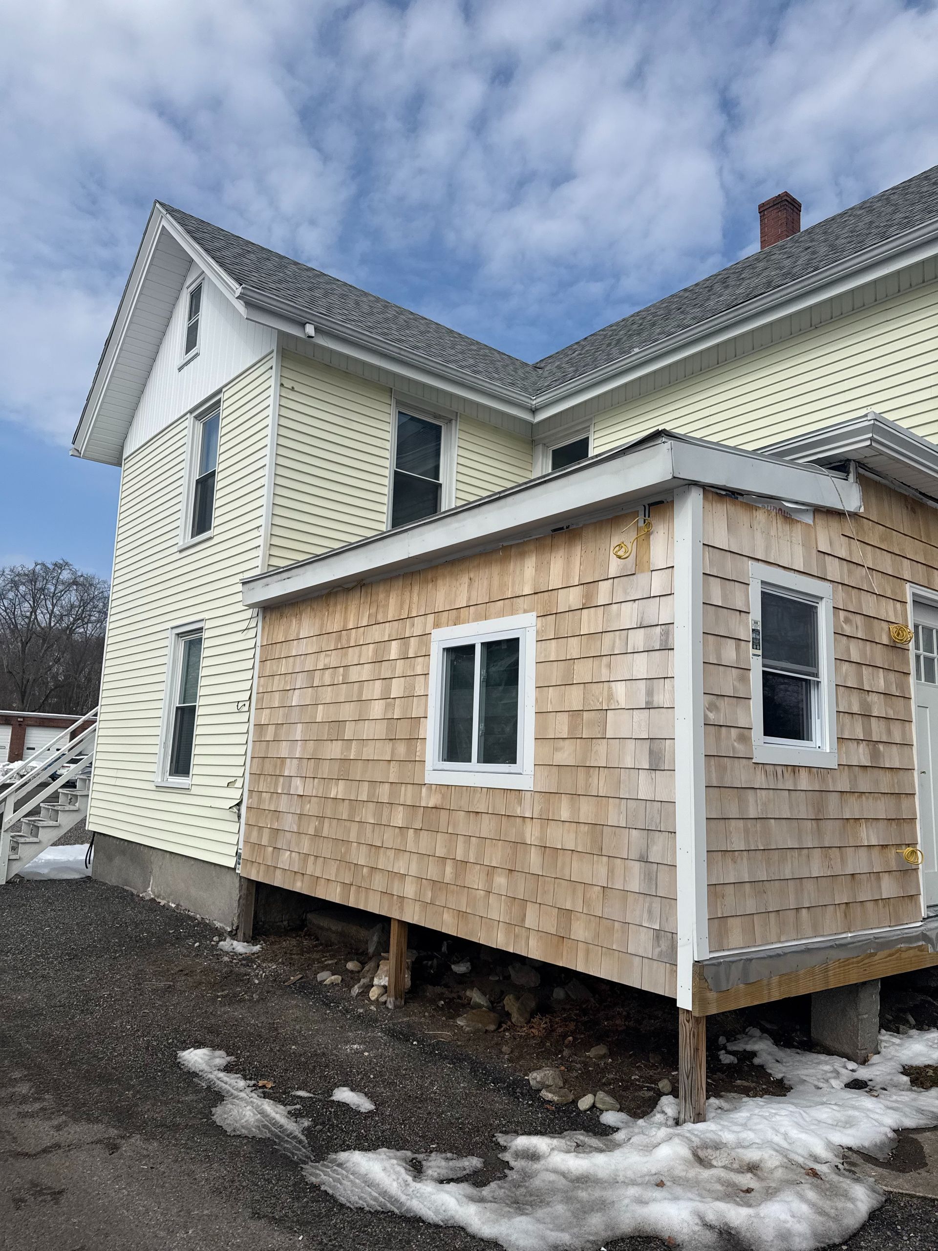 Yellow house with a wood-shingled addition; snow on the ground, cloudy sky.