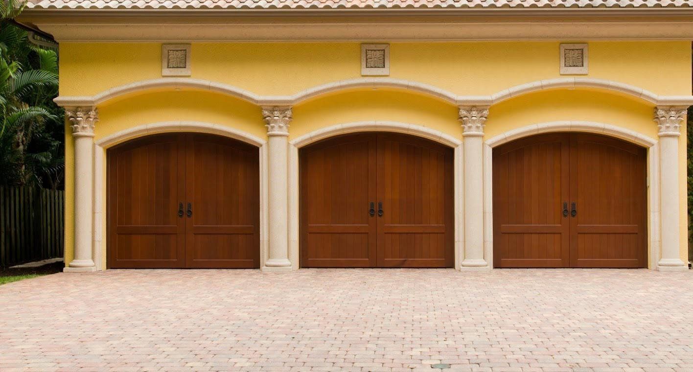 Three brown wooden garage doors under arched openings in a yellow building, with brick driveway.
