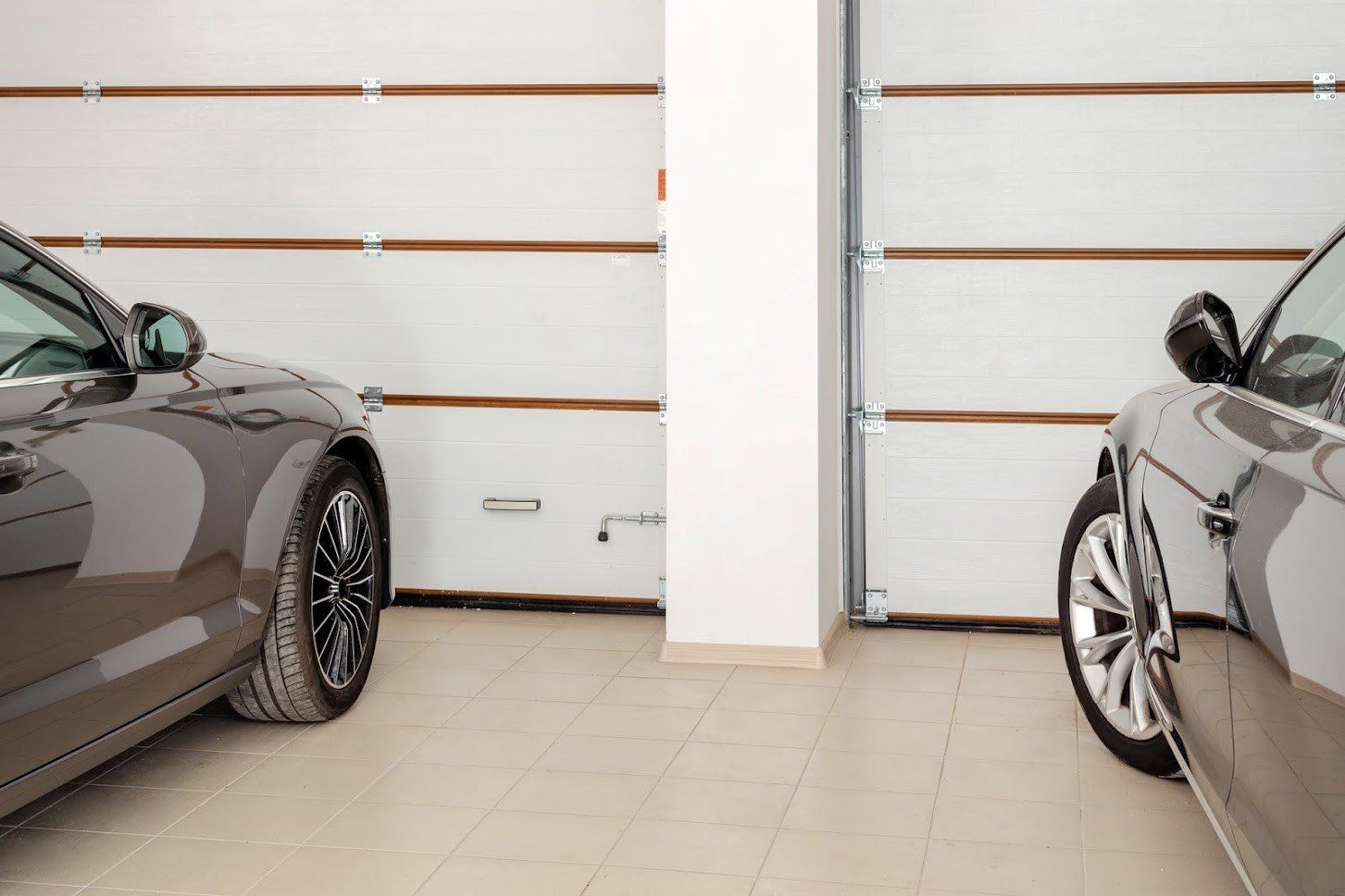 Two cars parked in a garage with closed white doors. A white pillar stands between them.