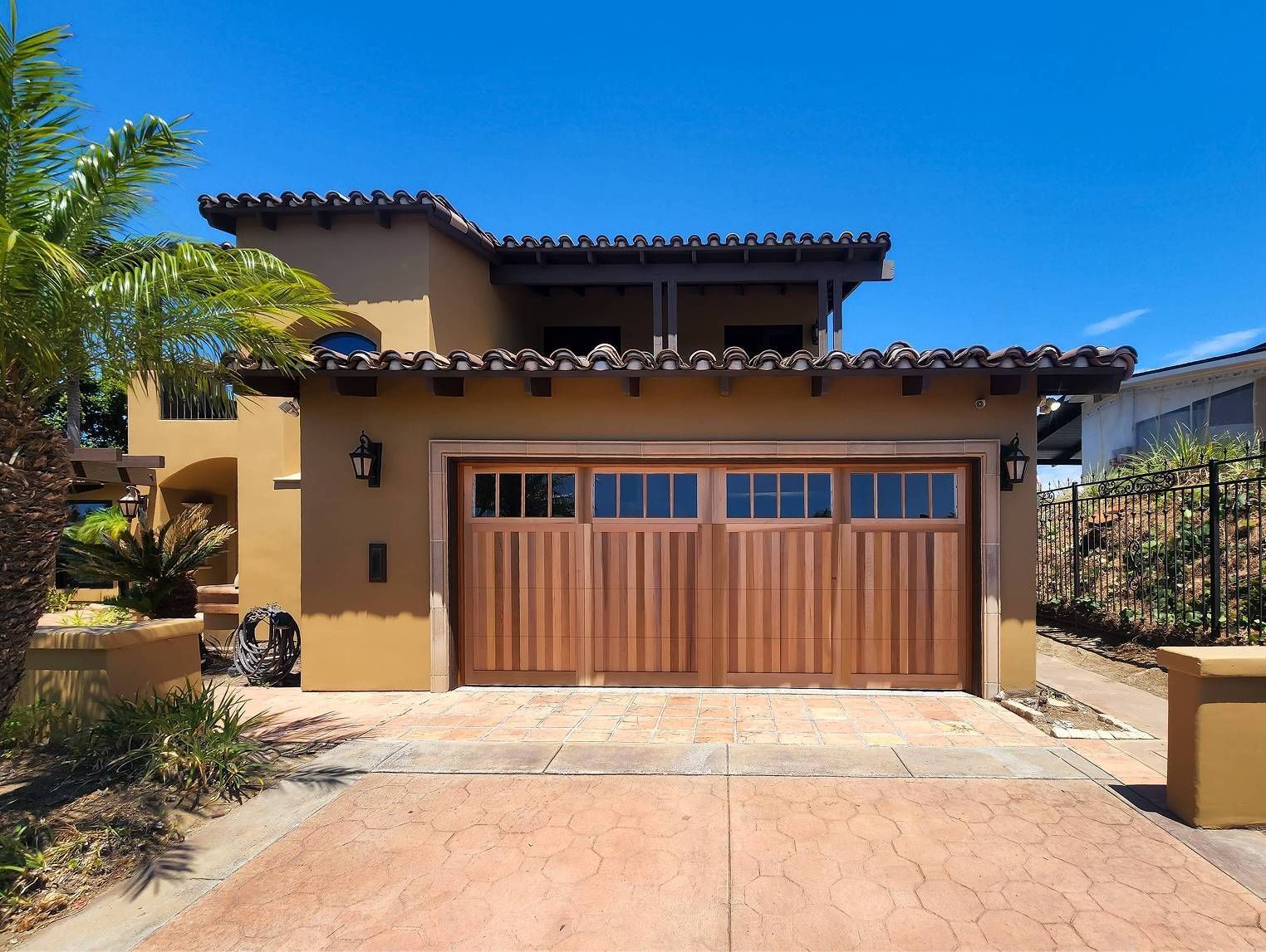 Two-story Spanish-style house with wooden garage door and terracotta roof under a bright blue sky.