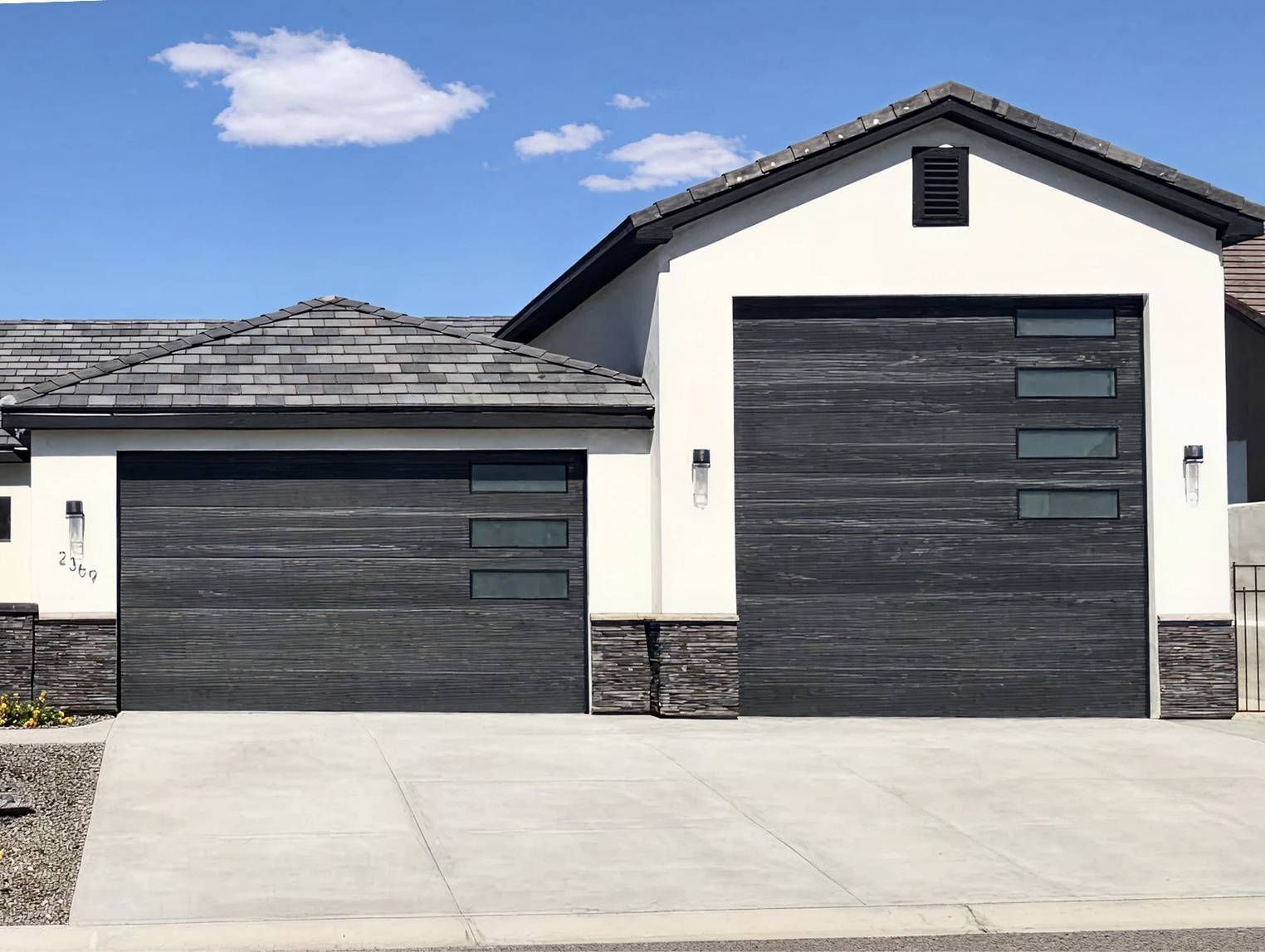 Modern home with two garage doors; dark wood-look with windows; light stucco walls, gray roof, and stone accents.