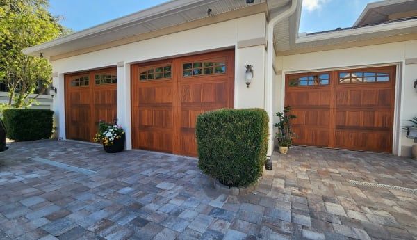 Three brown garage doors with glass panels, set in a paved driveway, surrounded by shrubs.