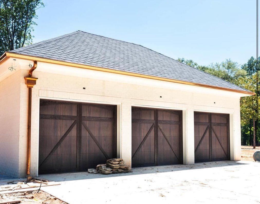 Three-car garage with dark wood doors, tan walls, and a gray shingled roof under a blue sky.