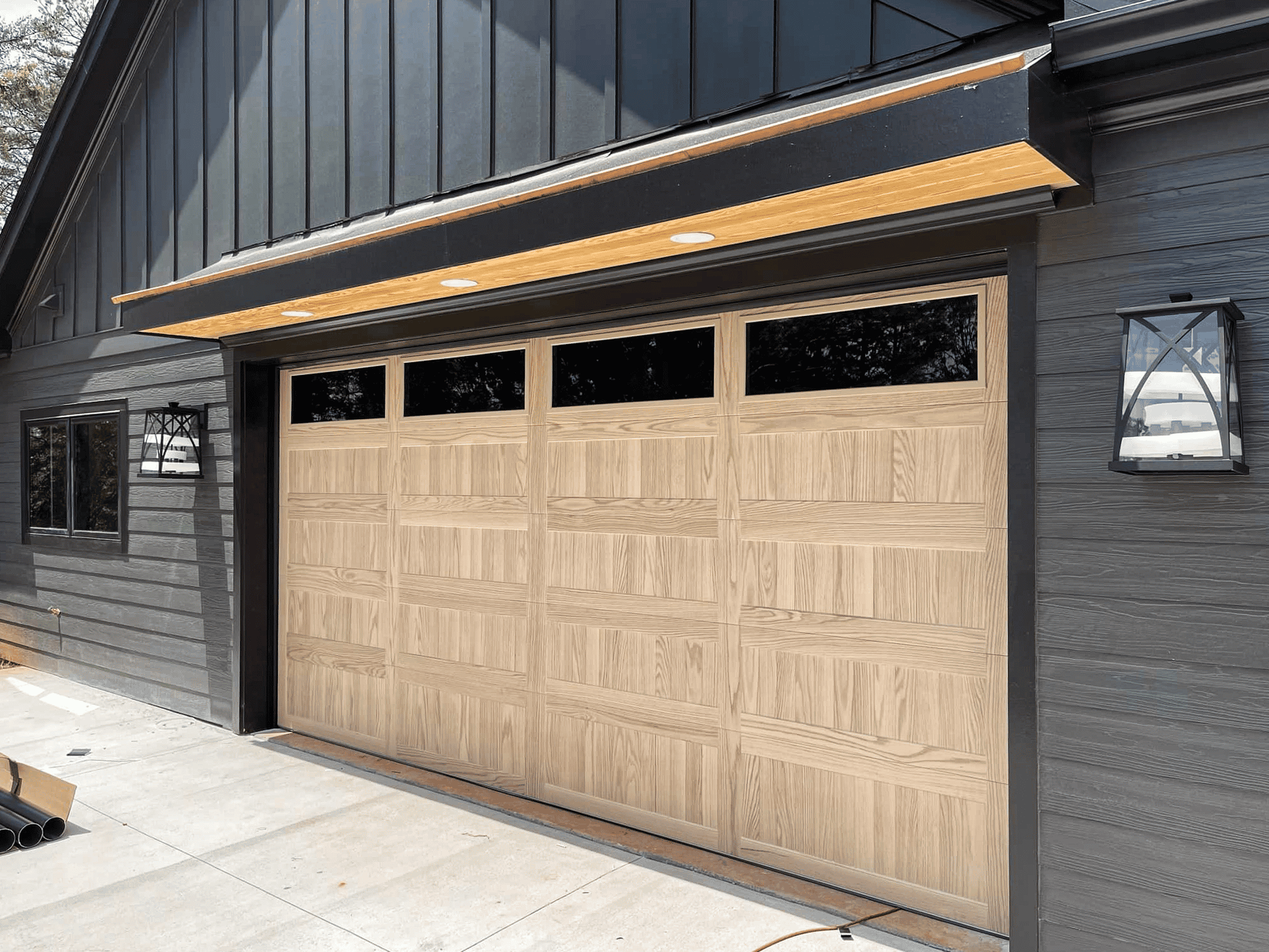Tan garage door with windows, under a black awning, next to a gray house.