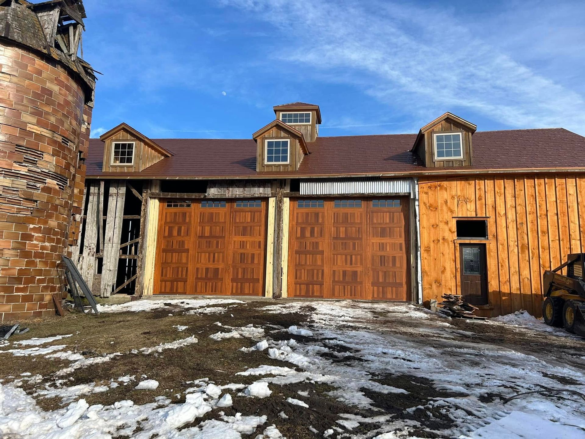 Rustic barn with brown garage doors, wooden siding, and a silo on a snowy day with a blue sky.