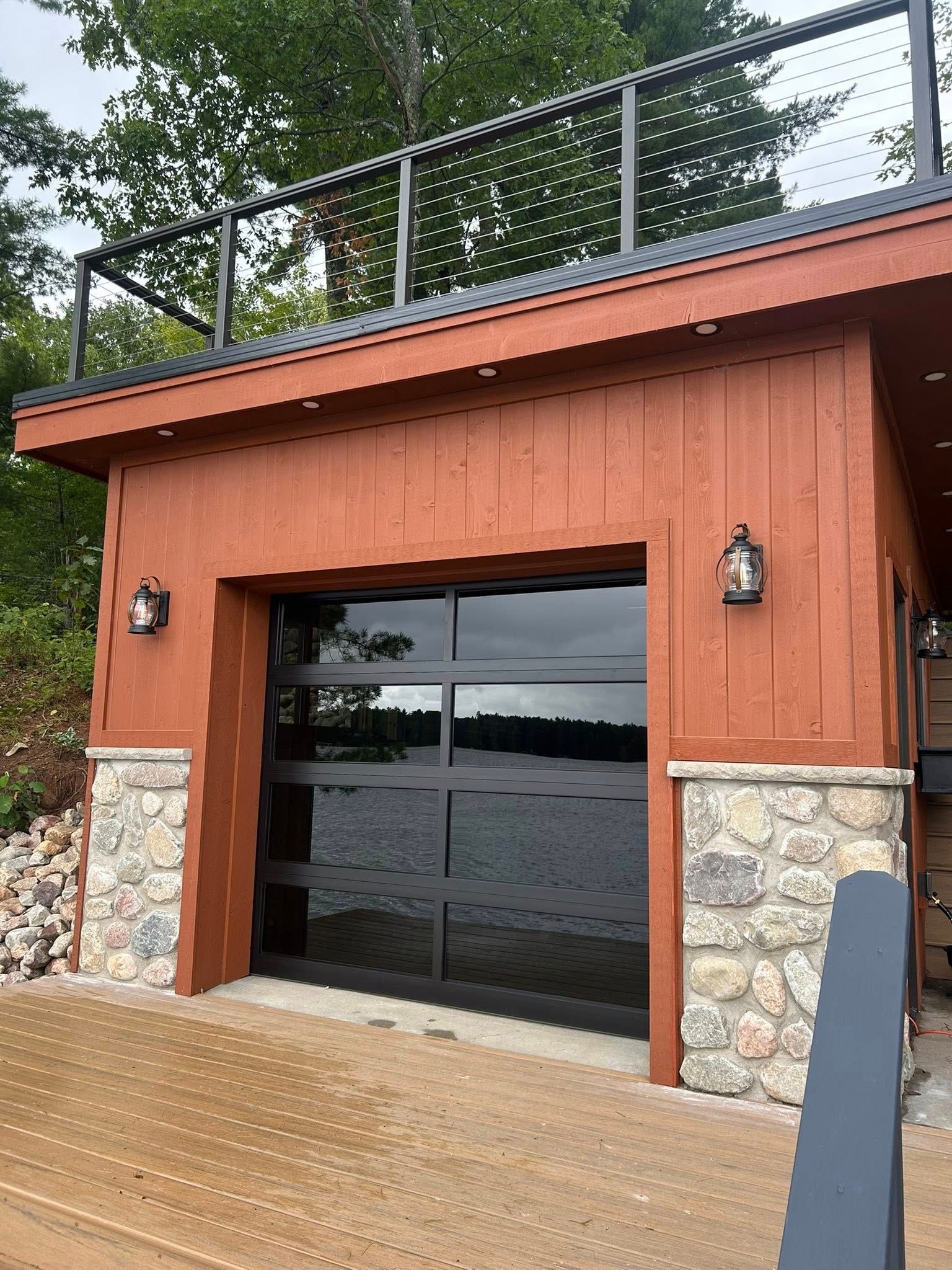 Red boathouse with glass garage door, stone accents, and rooftop deck overlooking water.