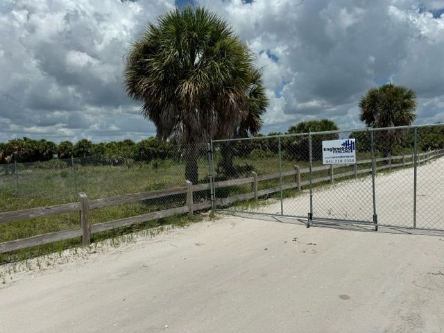 A chain link fence surrounds a dirt road with palm trees in the background.