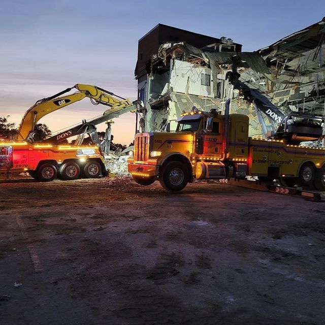 Two tow trucks with lights working to dismantle a building at dusk. Yellow truck, excavator.
