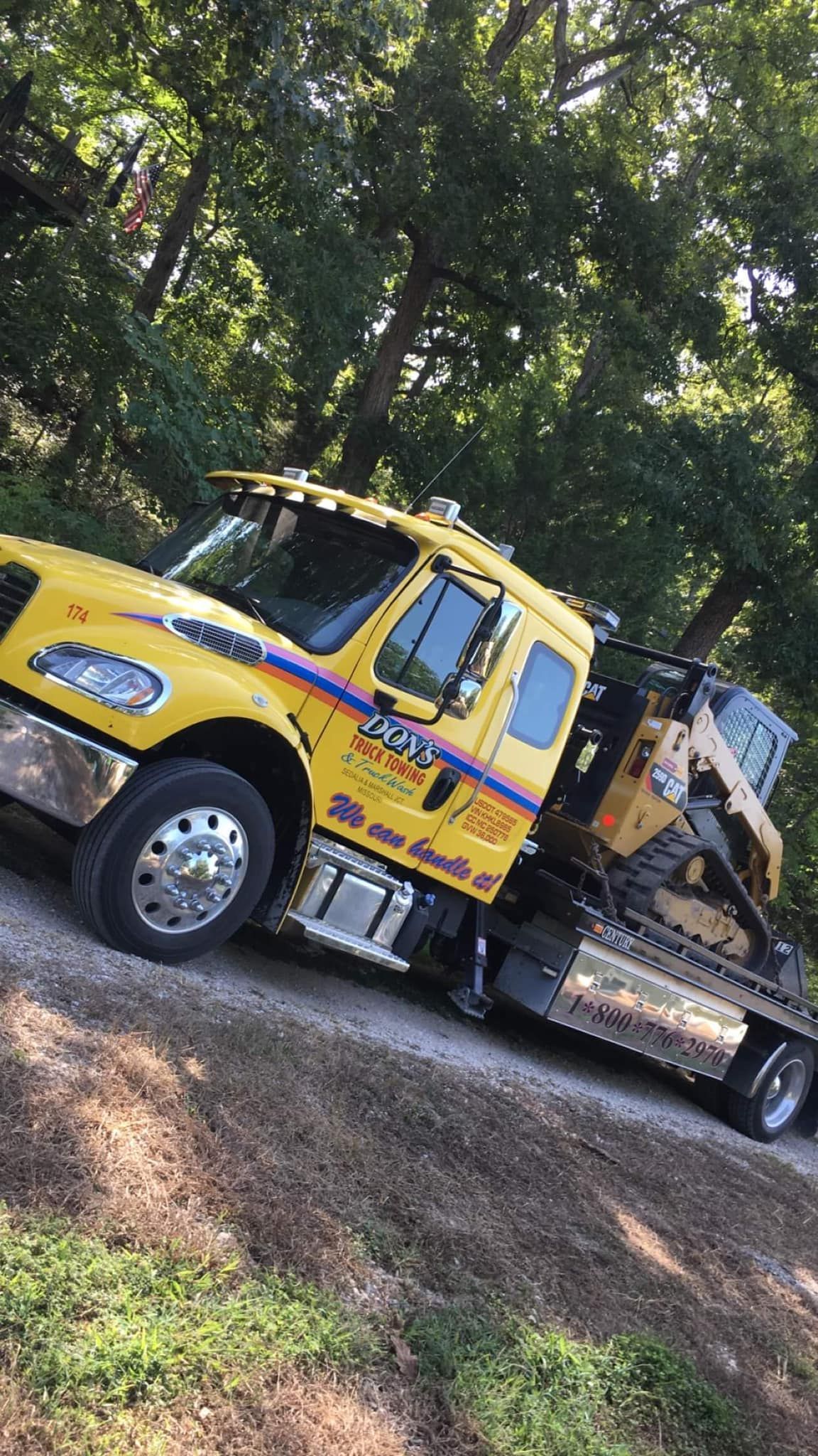 Yellow tow truck hauling construction equipment on a dirt road, surrounded by trees.
