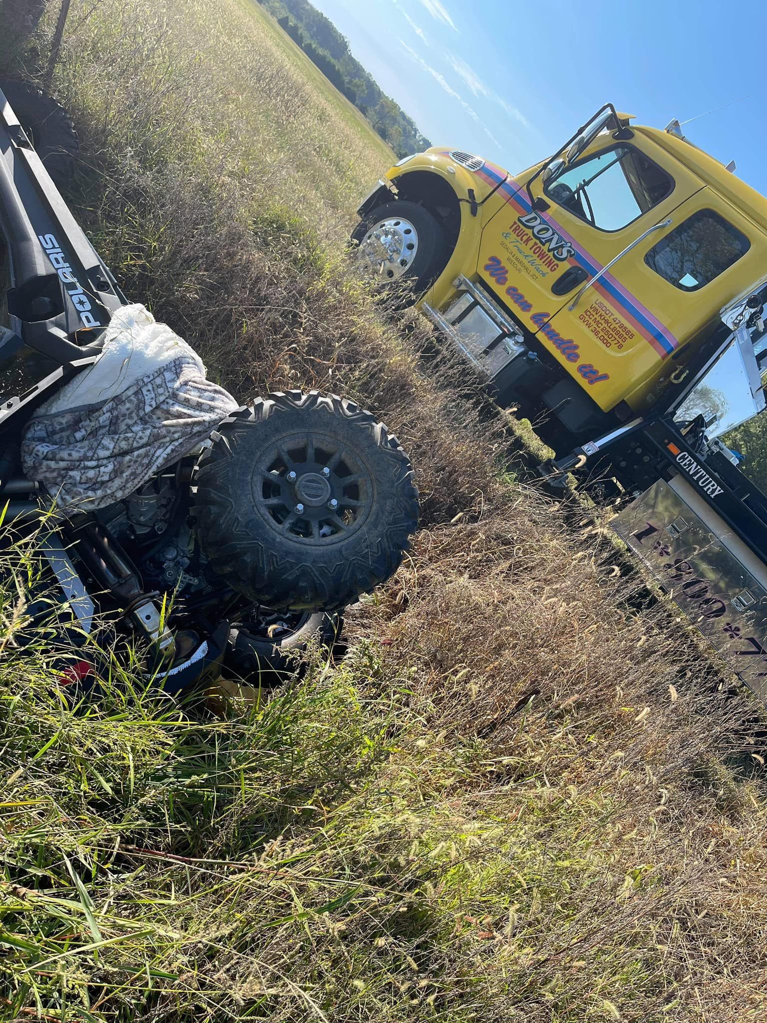 Yellow tow truck in a field of tall dry grass, partially overturned, with a blue sky.