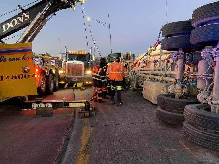 A flipped semi-truck being recovered by a tow truck at a roadside scene; emergency responders assess.