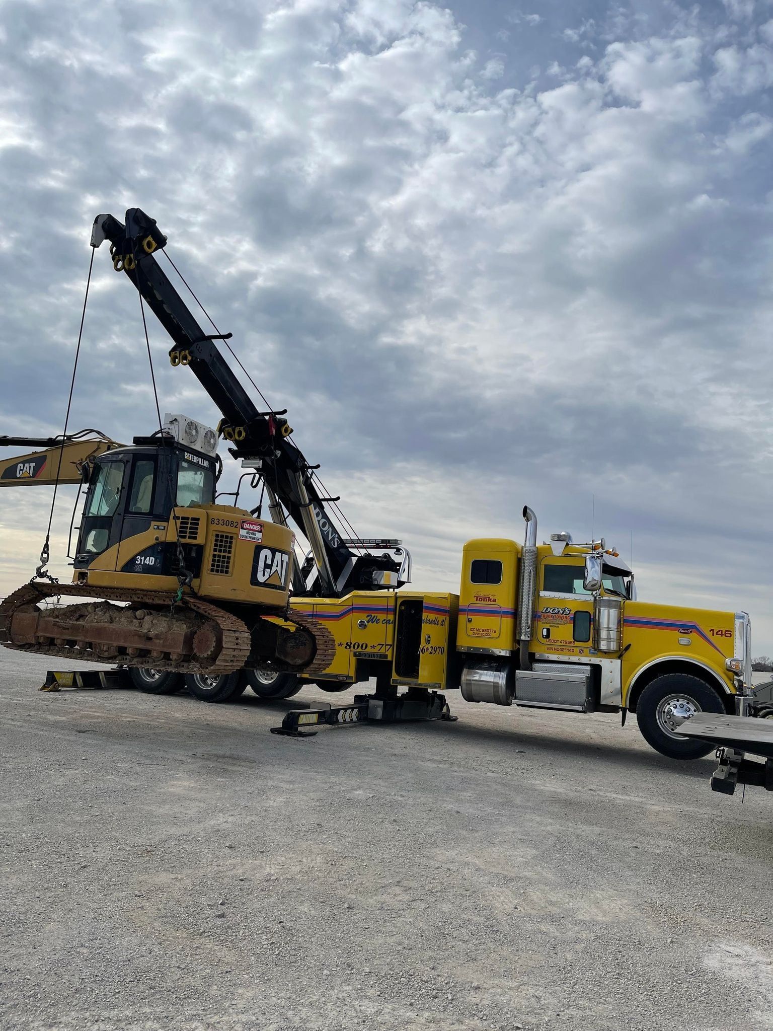 Yellow tow truck lifting a yellow Caterpillar bulldozer on a gravel lot under a cloudy sky.