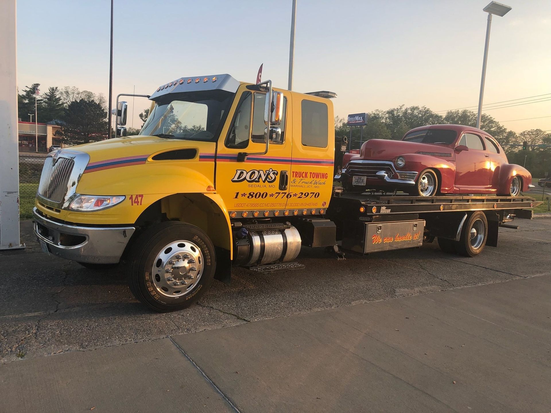 Yellow tow truck carrying a red classic car on a flatbed in a setting.