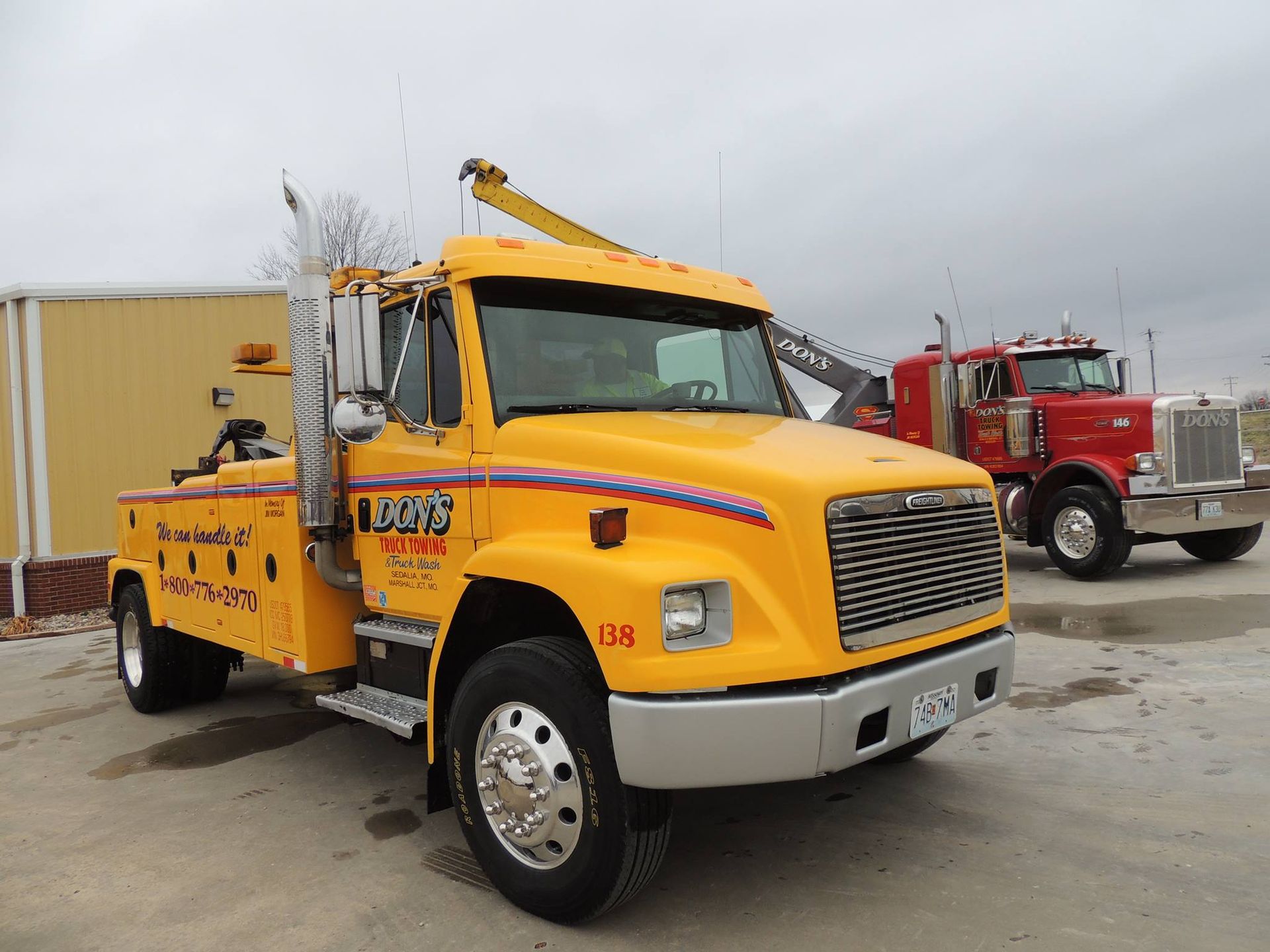 Yellow tow truck parked, red semi-truck in the background. Cloudy sky, outdoor setting.