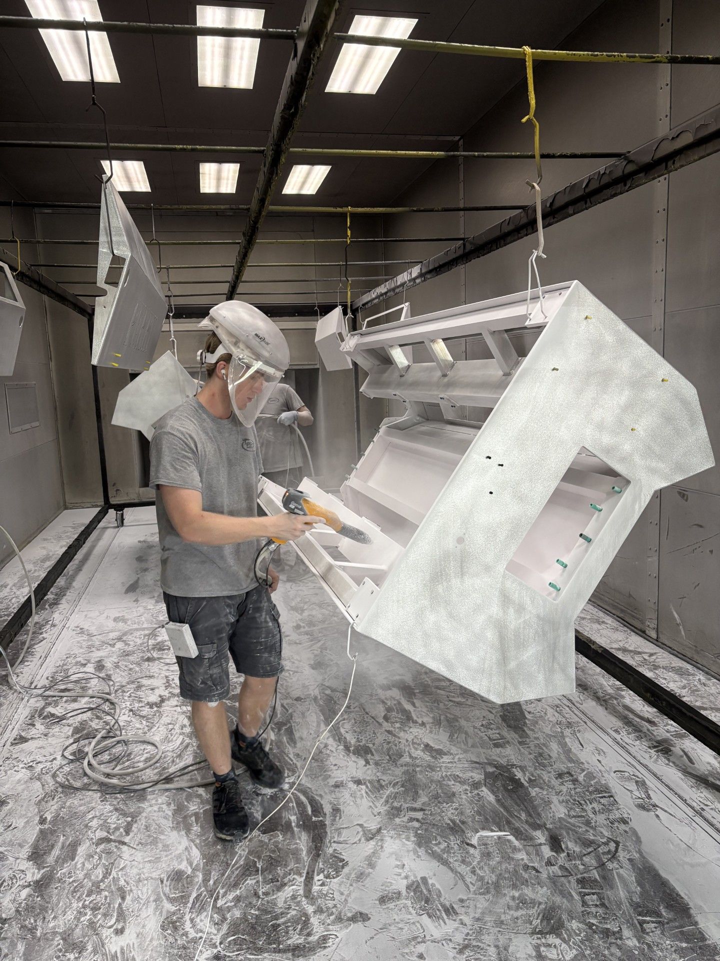 Man powder coating metal parts in a spray booth, wearing protective gear.