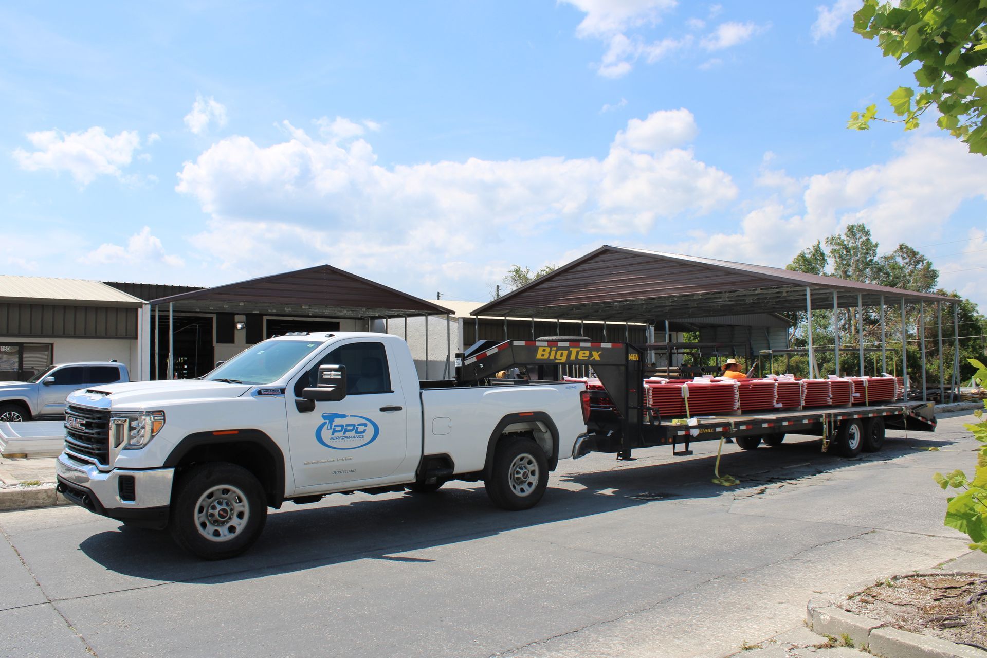 White truck with trailer hauling building materials on a street in front of a building on a sunny day.