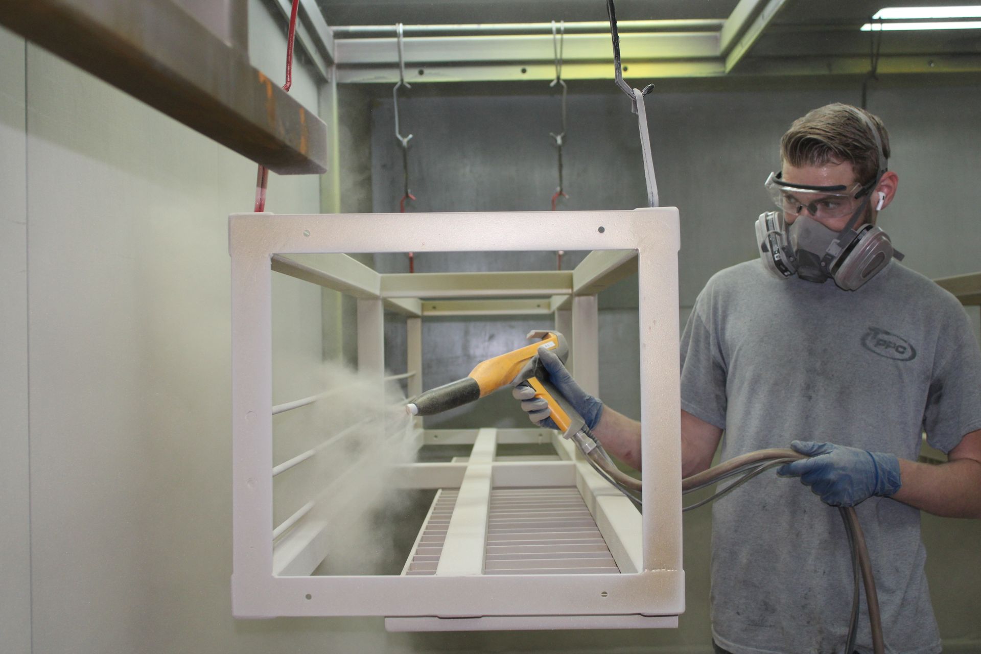 A worker spraying powder coating on a wooden frame, wearing safety glasses and a respirator in a workshop.