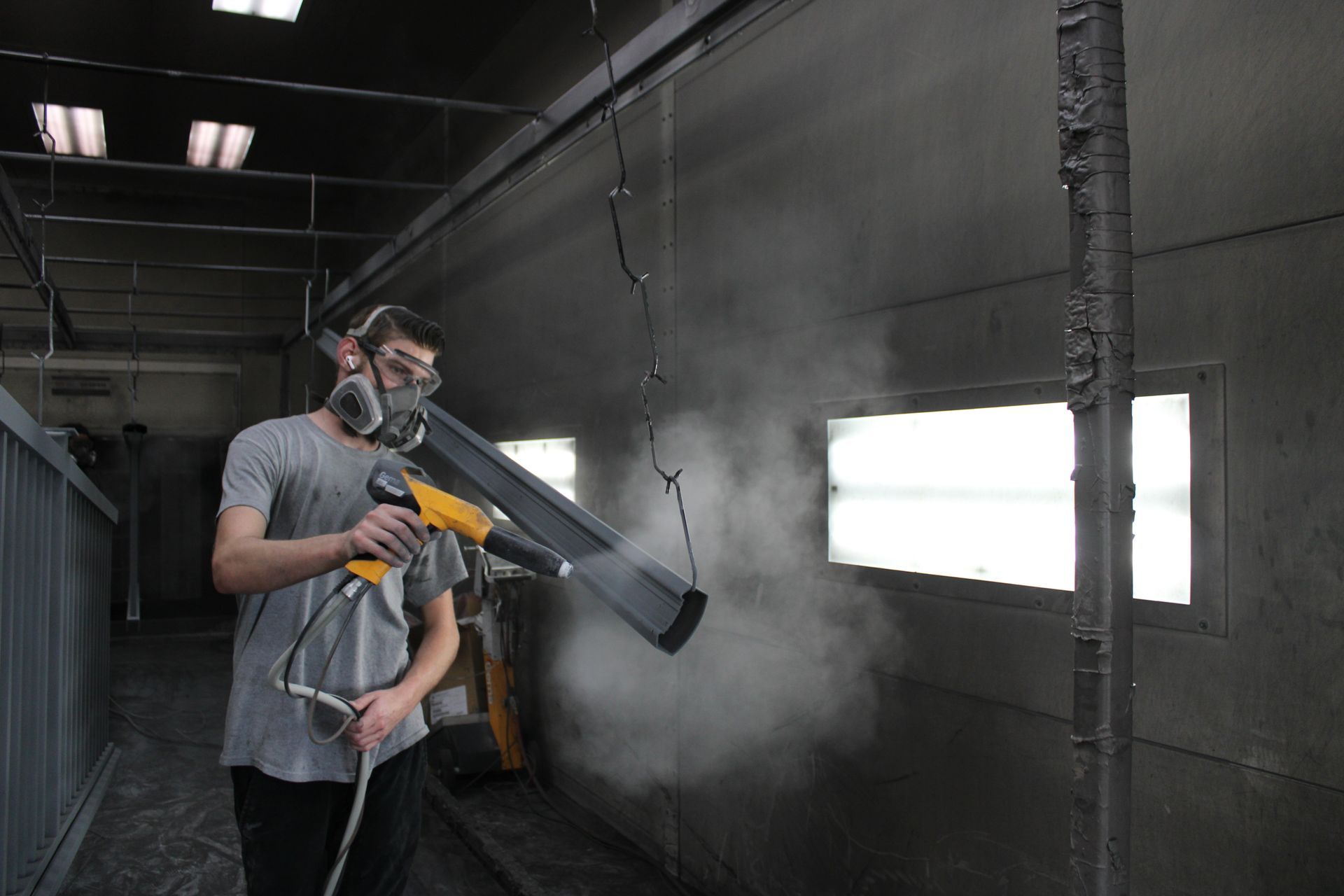 Man spraying powder coat in a booth. He wears a respirator, goggles, and holds a spray gun.