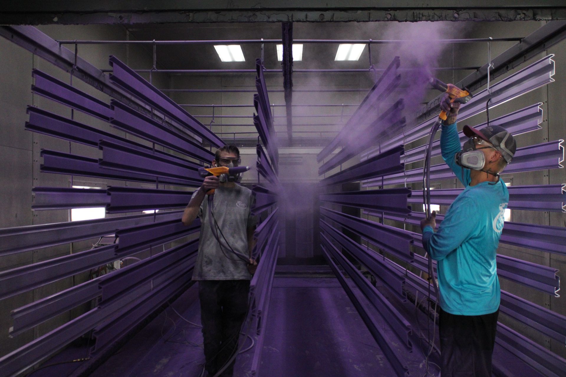 Two workers spray purple paint on metal beams in an industrial setting.