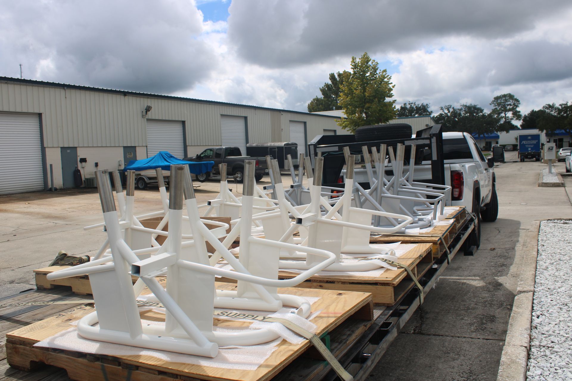 White exercise equipment on pallets loaded on a truck bed, outside a factory on a cloudy day.