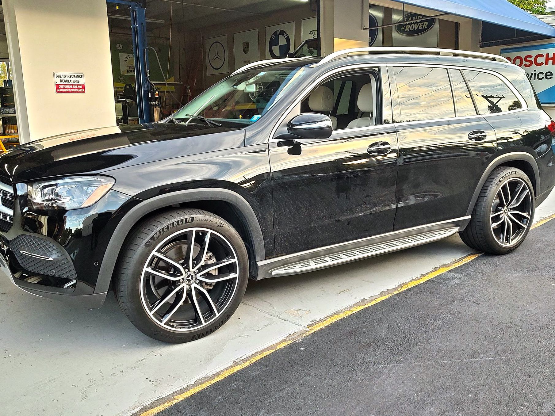 Black Mercedes-Benz GLS SUV parked in front of a repair shop, with custom black and silver wheels.
