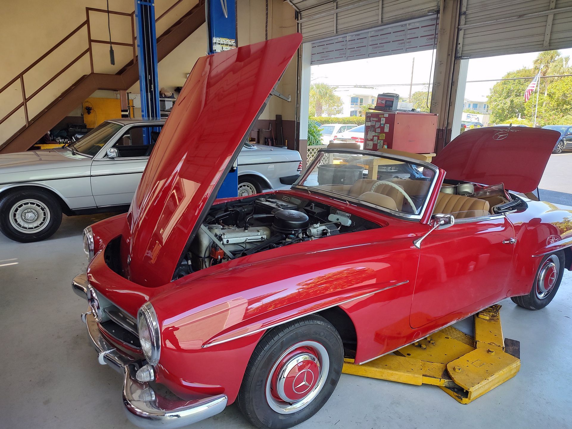 Red classic Mercedes-Benz convertible with open hood, on a lift in a garage. Another white car is nearby.