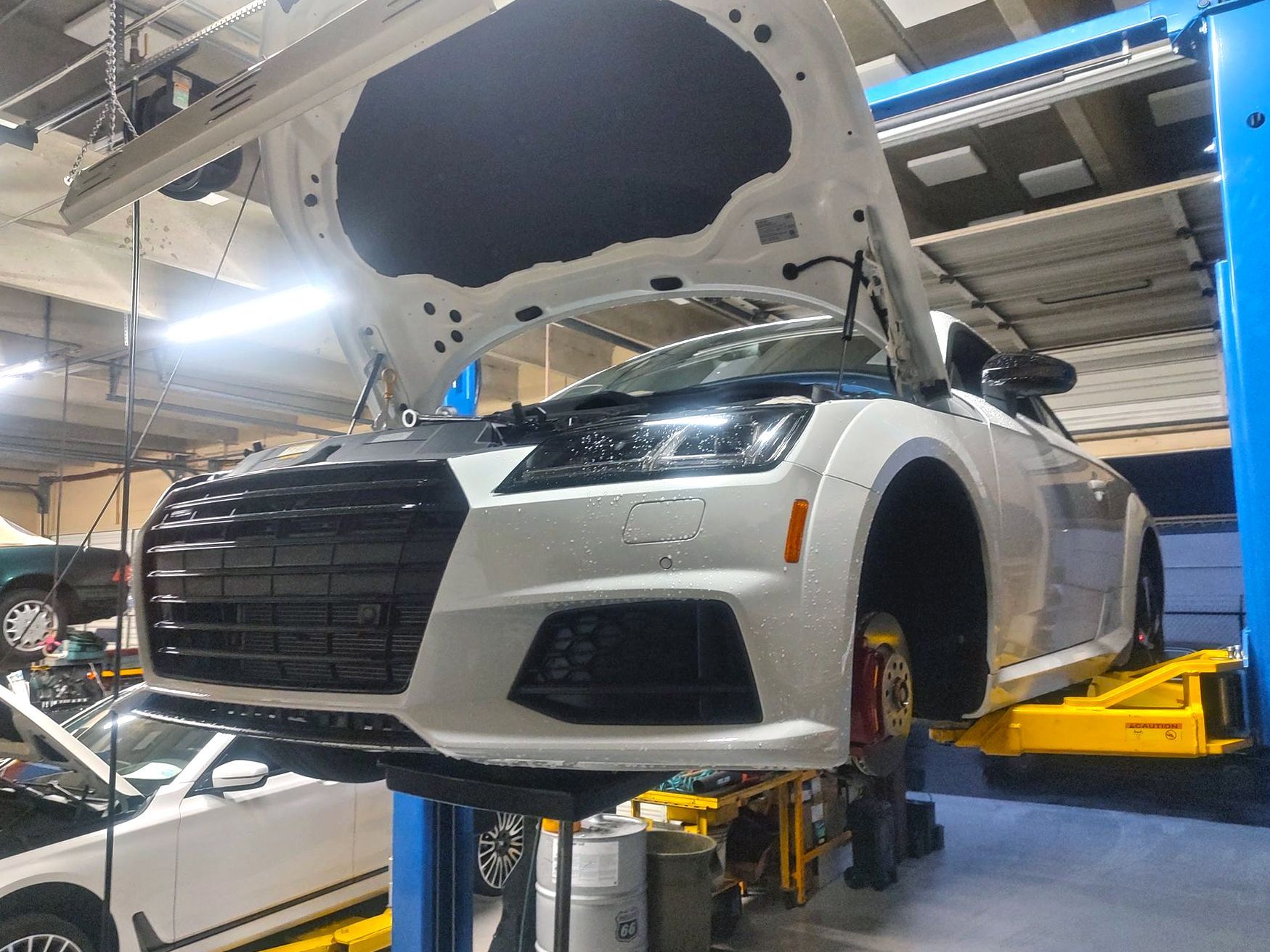 White Audi TT on a car lift with hood open in an auto repair shop.