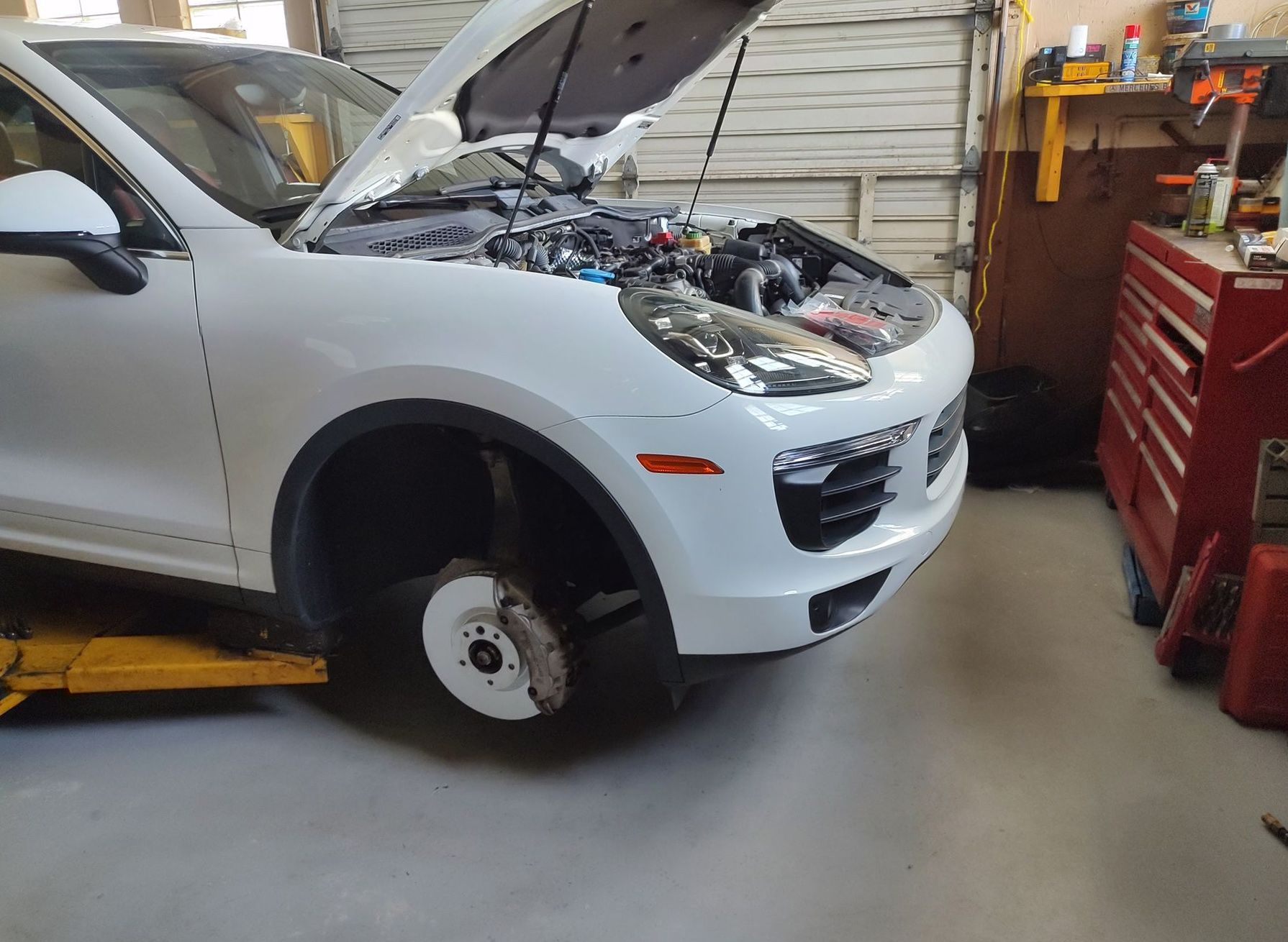 White car with hood up, front wheel removed, in a garage.
