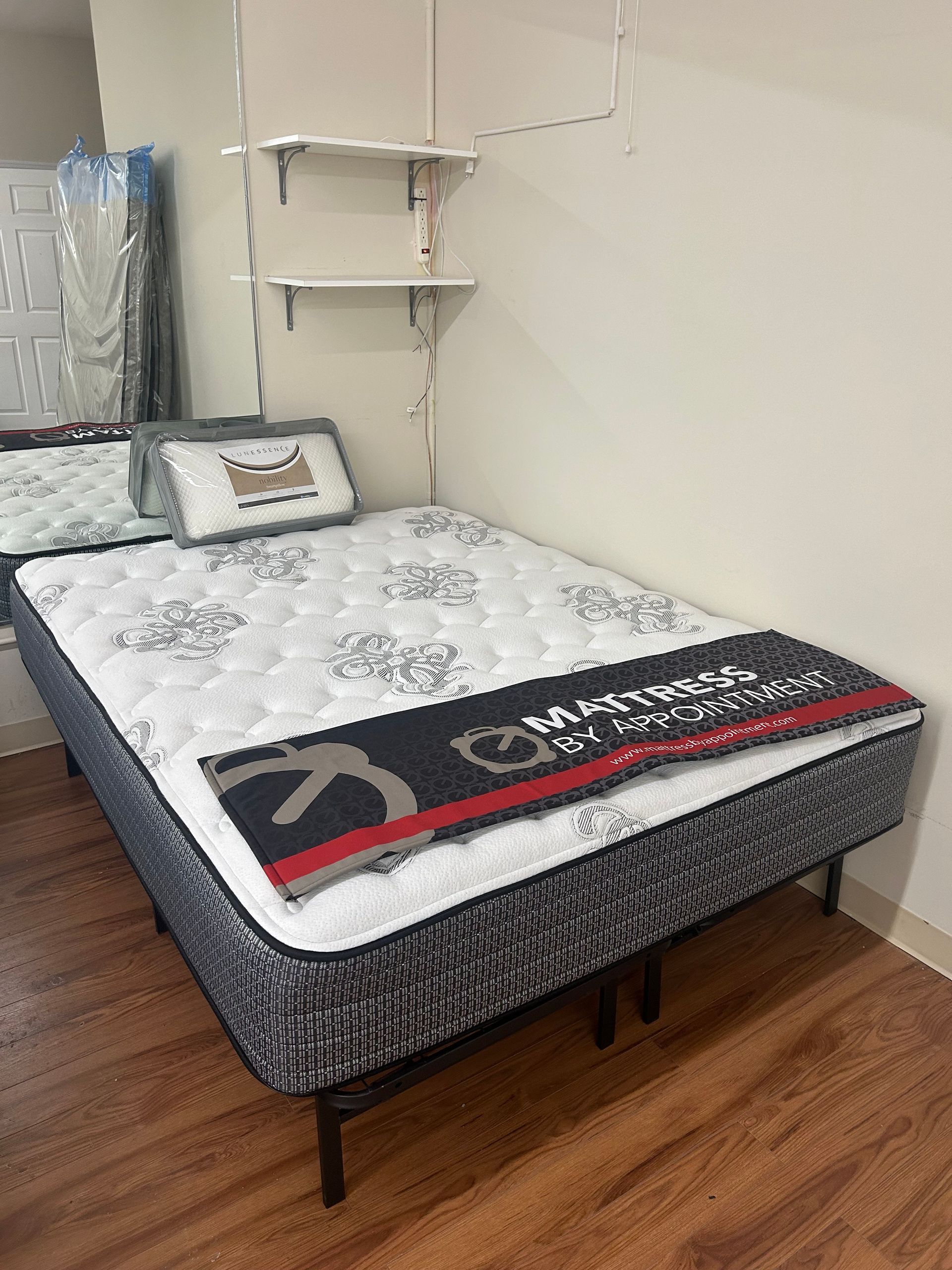 Bed with mattress, pillows, and a blanket in a room with white walls, and wooden floors