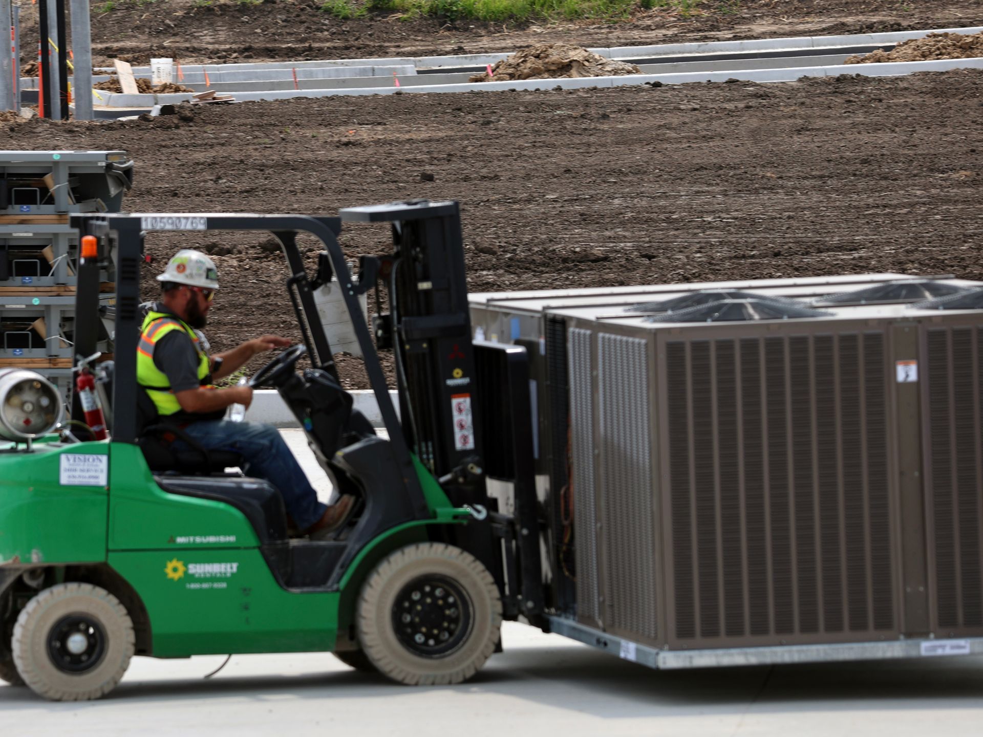 A man is driving a green forklift carrying a large air conditioner.
