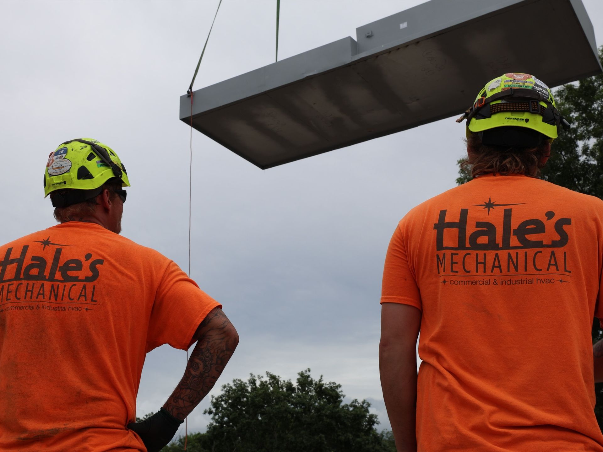 Two men wearing orange Hale's Mechanical shirts and safety helmets watching a large metal panel being lifted.