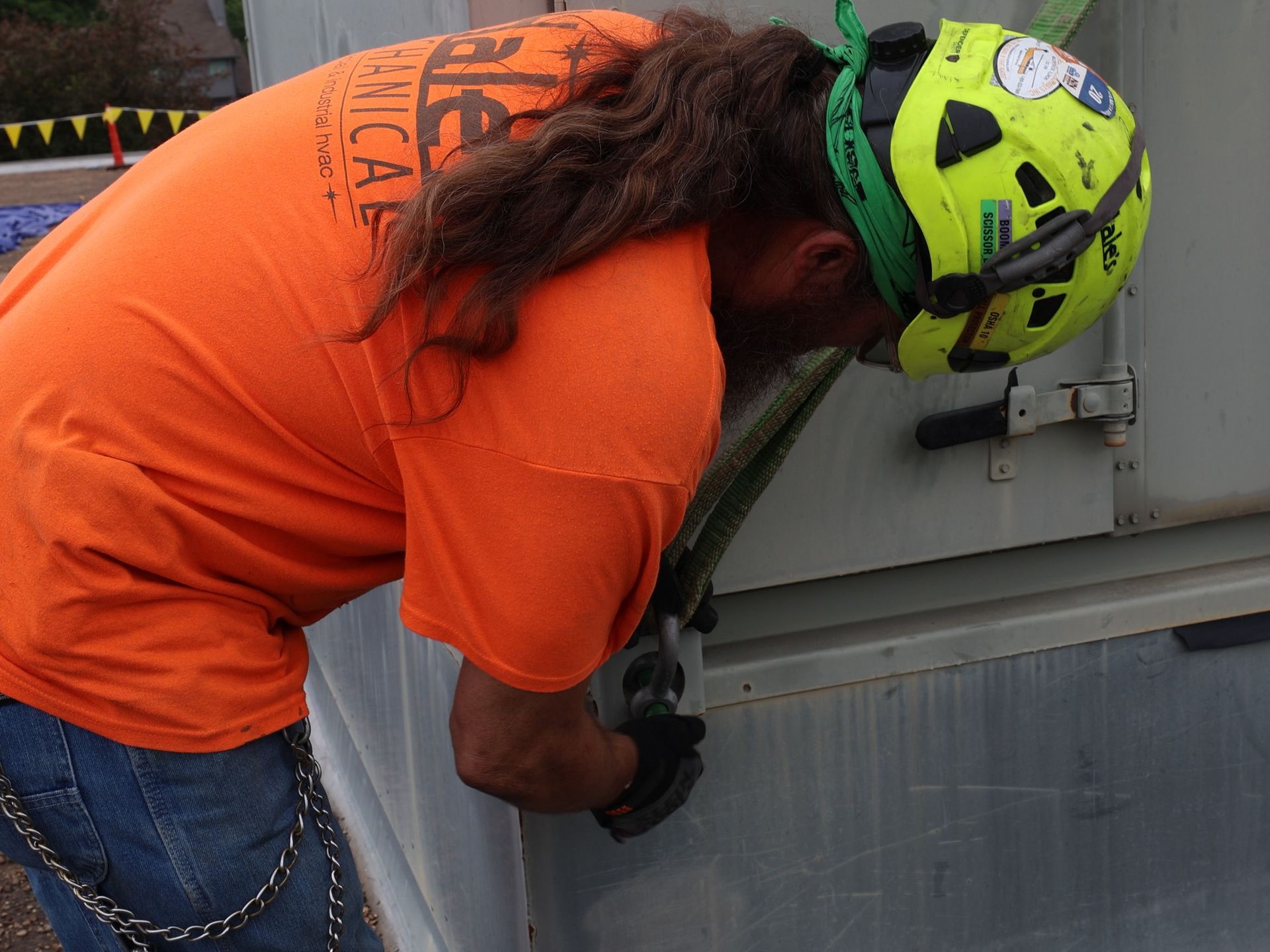 Man in orange shirt and hard hat, working on a gray container, outdoors.