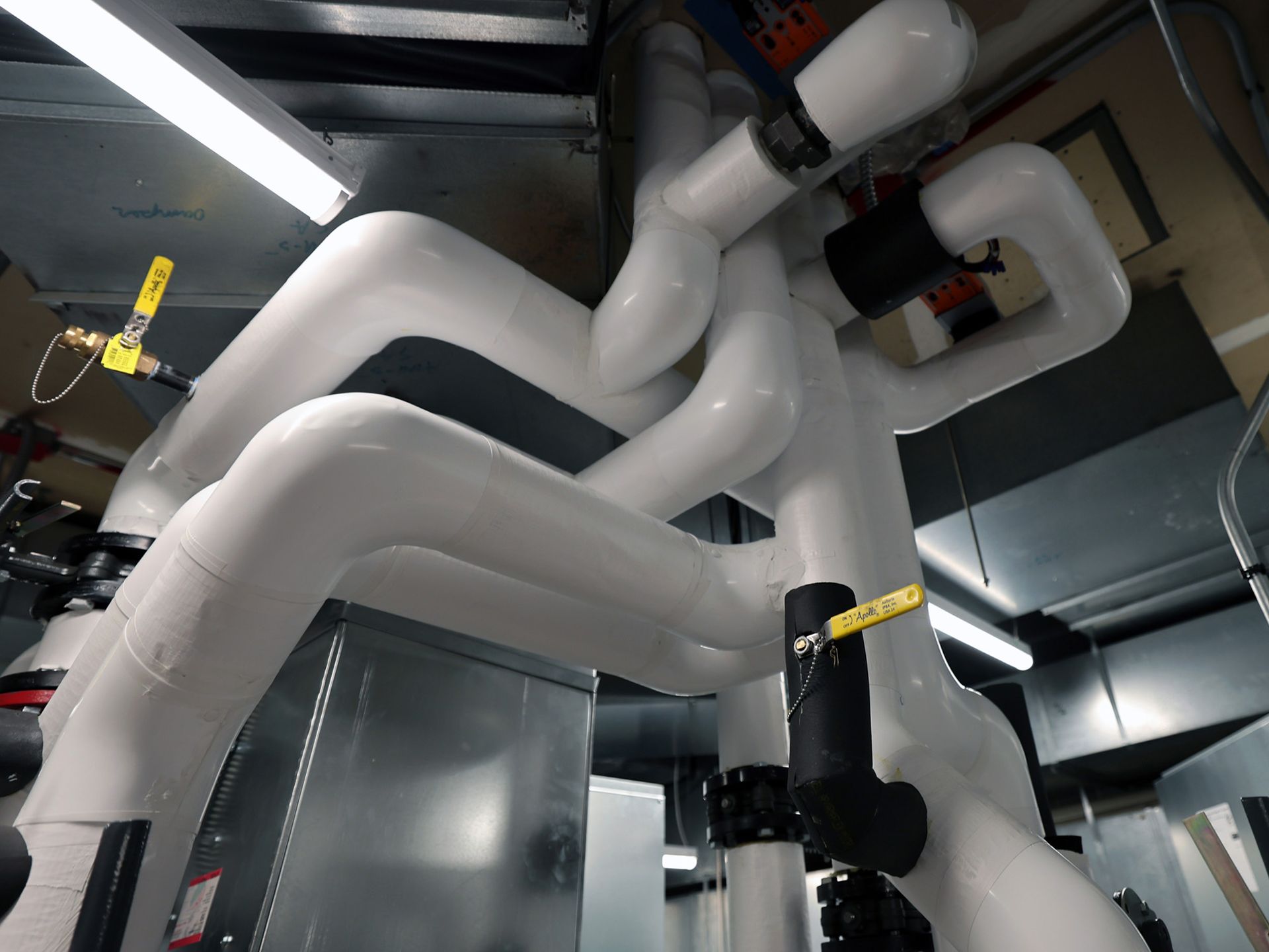 White insulated pipes in a mechanical room with yellow valve handles and fluorescent lights.