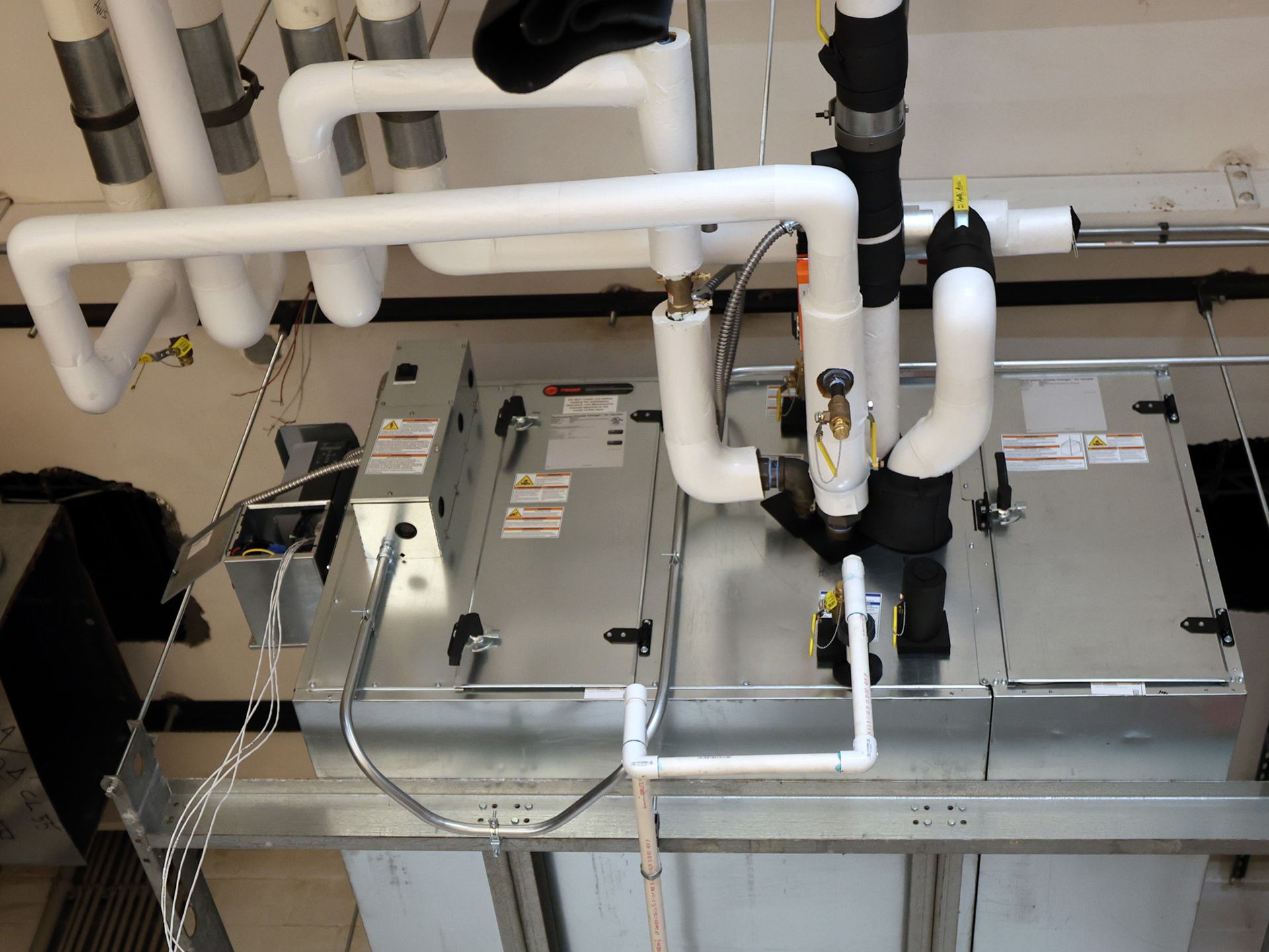 HVAC unit with insulated pipes, electrical wiring, and labels, mounted on a ceiling.