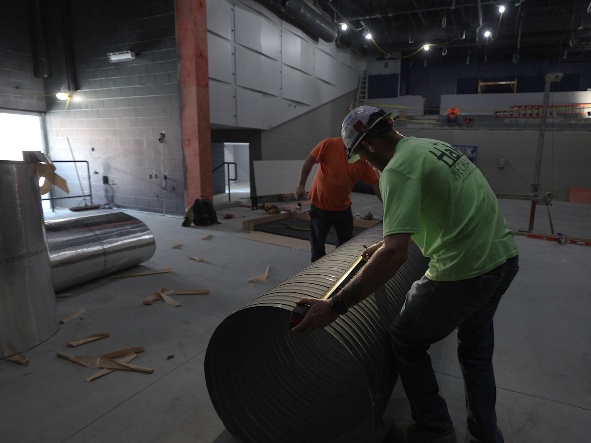 Two construction workers installing a large, corrugated metal duct in a building with exposed walls and ceilings.