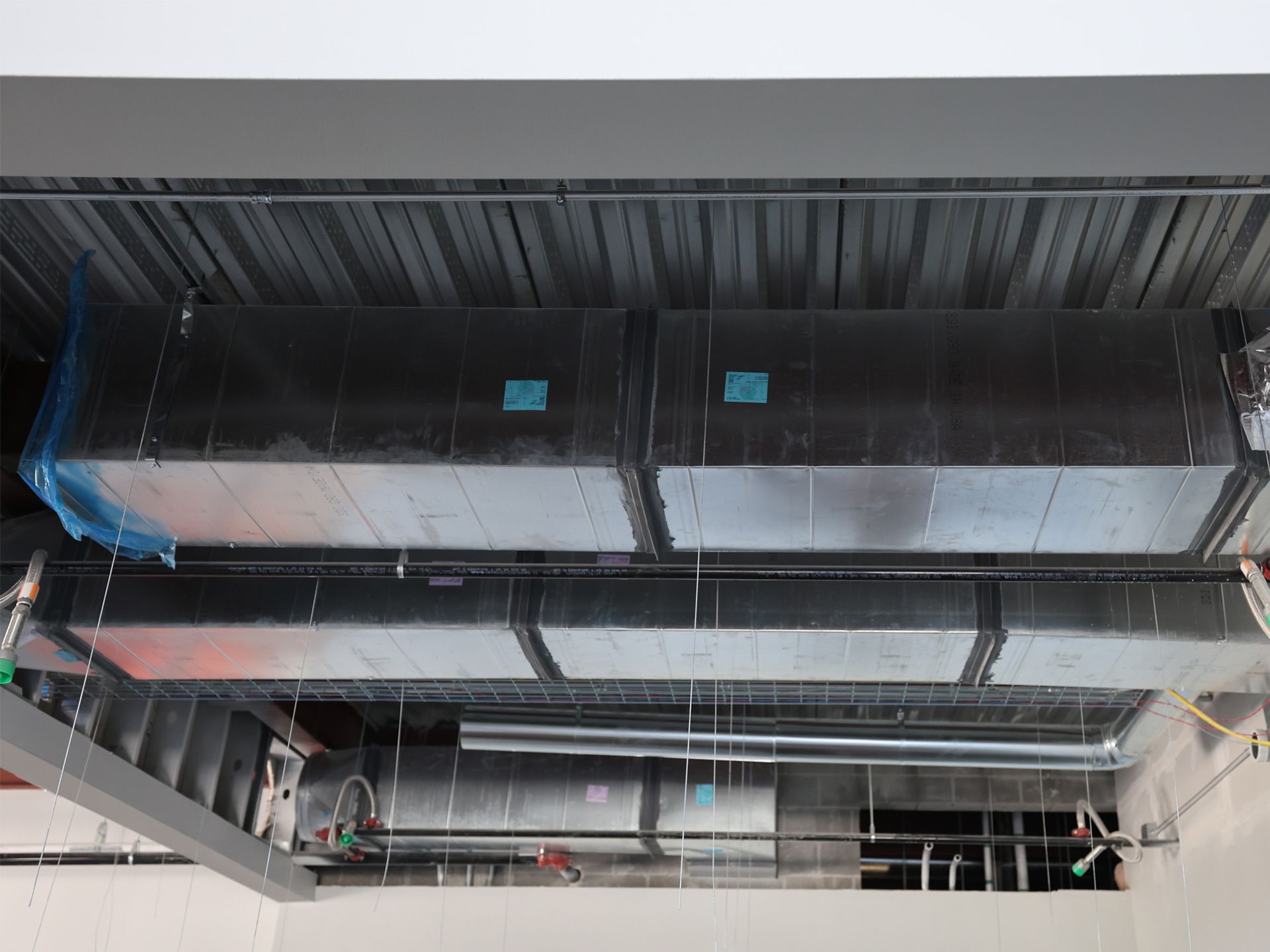 Air ducts installed in a building, viewed from below. Rectangular, silver ducts against gray ceiling, with blue tape and protective cover.