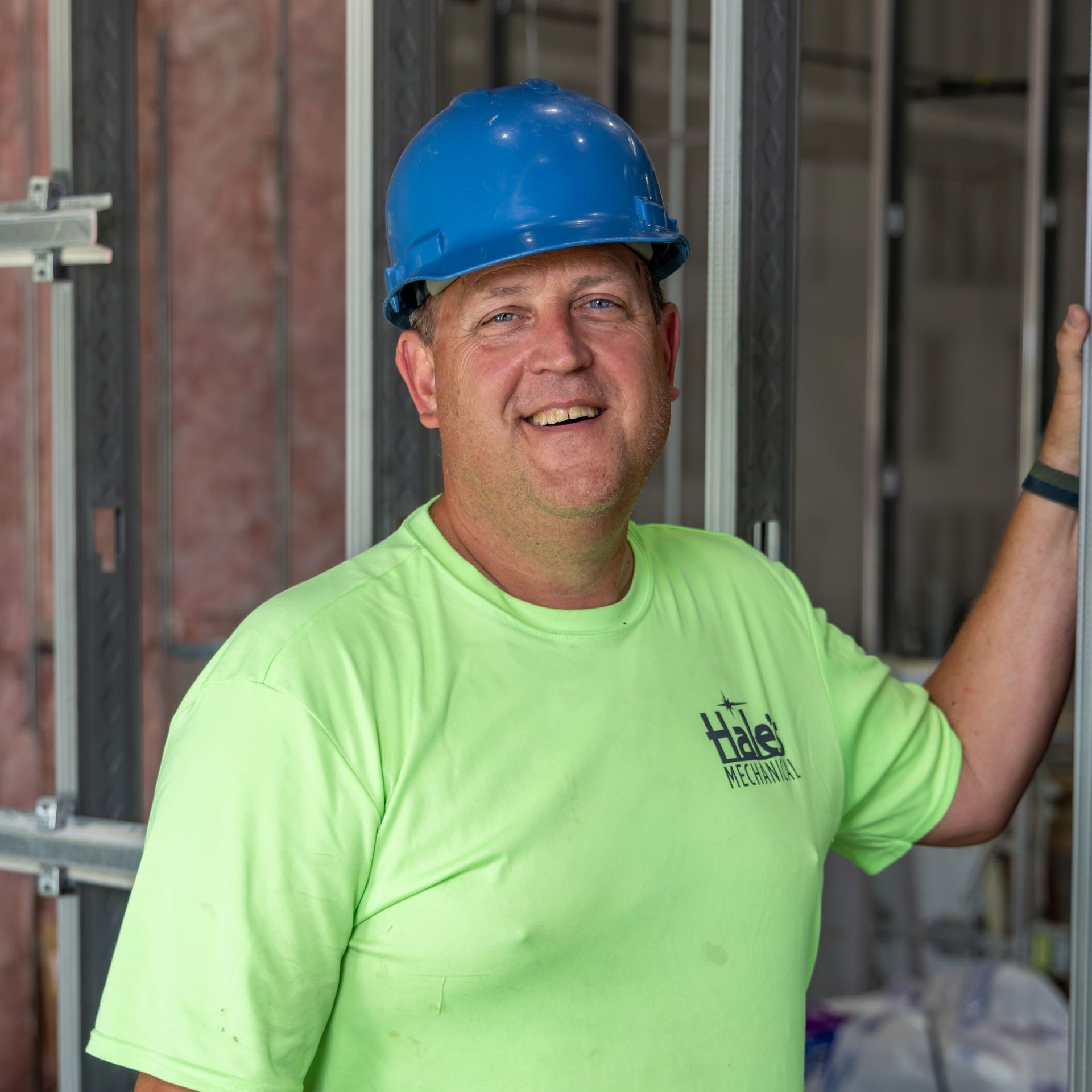 A man wearing a blue hard hat and a green shirt is leaning against a wall.