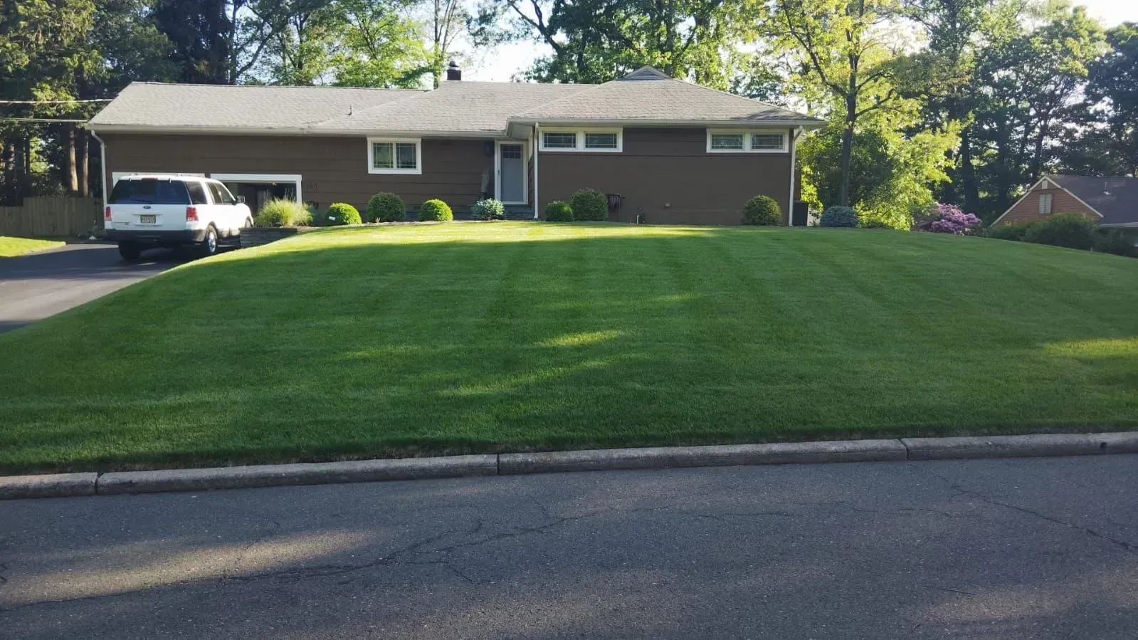 Brown house with a well-manicured green lawn sloping down to the street. A white car is parked in the driveway.
